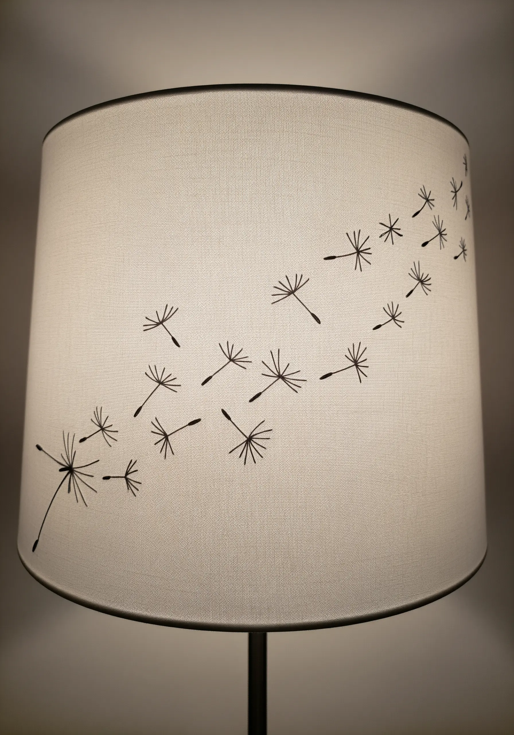 A white drum lampshade embroidered with a delicate design of dandelion seeds blowing in the wind.