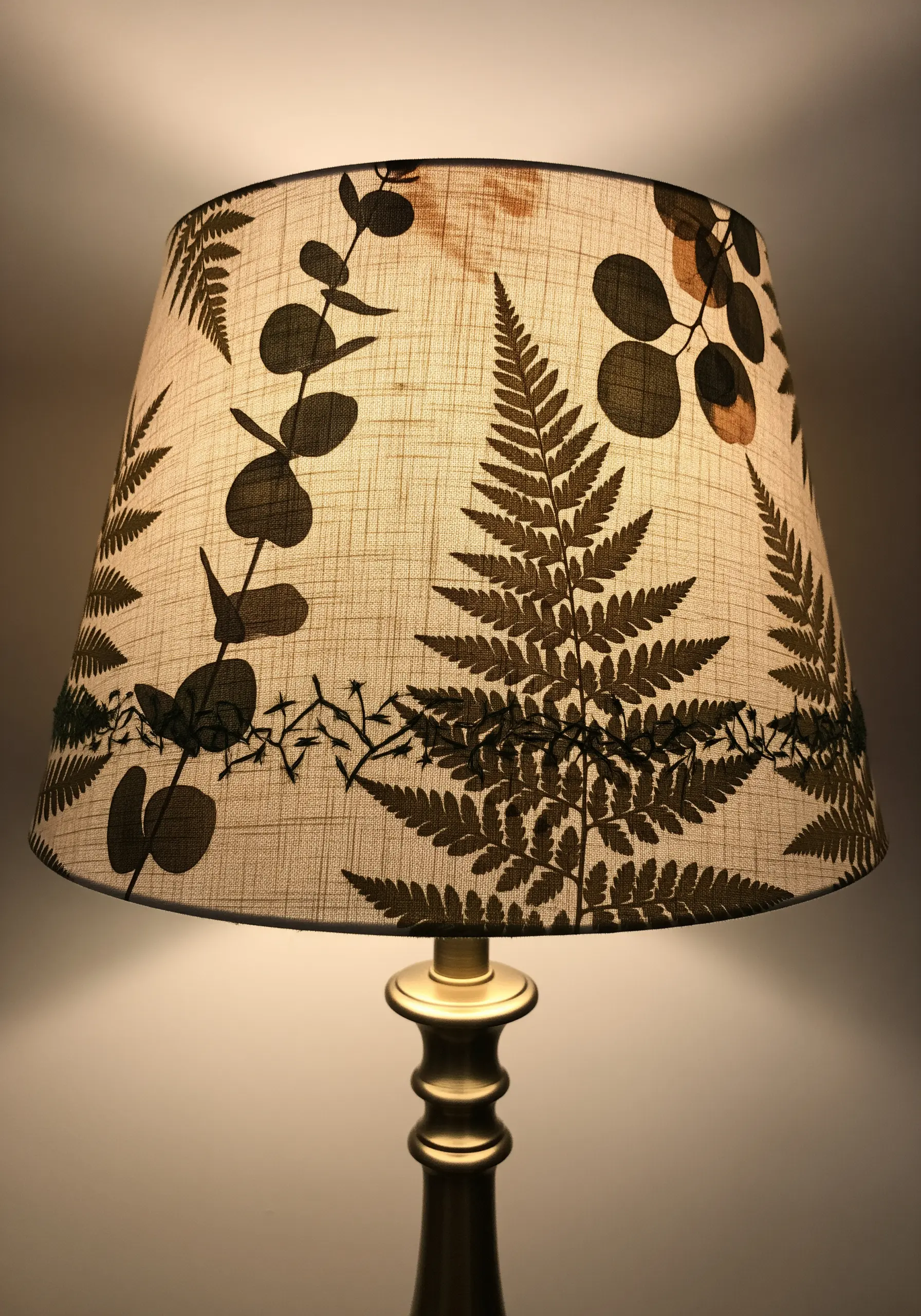 A linen lampshade embroidered with dark brown silhouettes of fern and eucalyptus leaves.