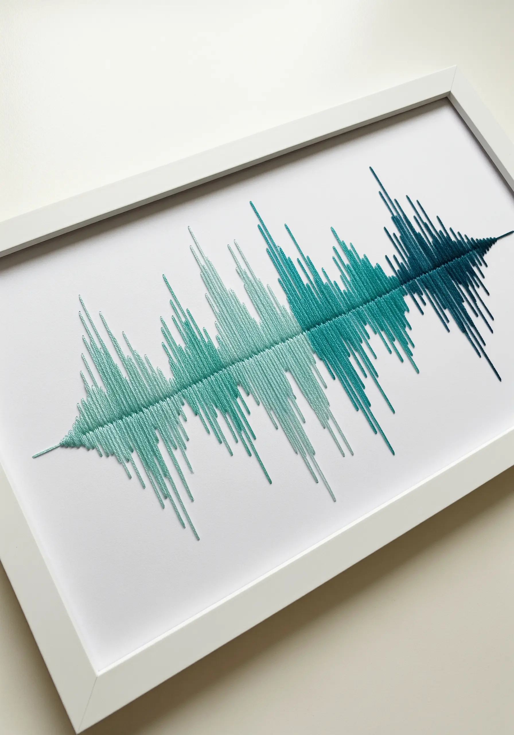 An embroidered soundwave in shades of green and teal on white paper, displayed in a white frame.