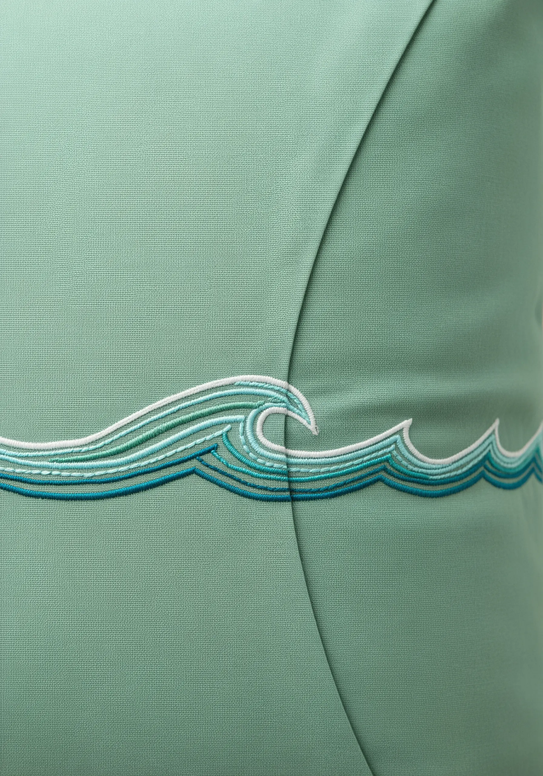 A stylized ocean wave embroidered in shades of blue, teal, and white on a seafoam green pillow.