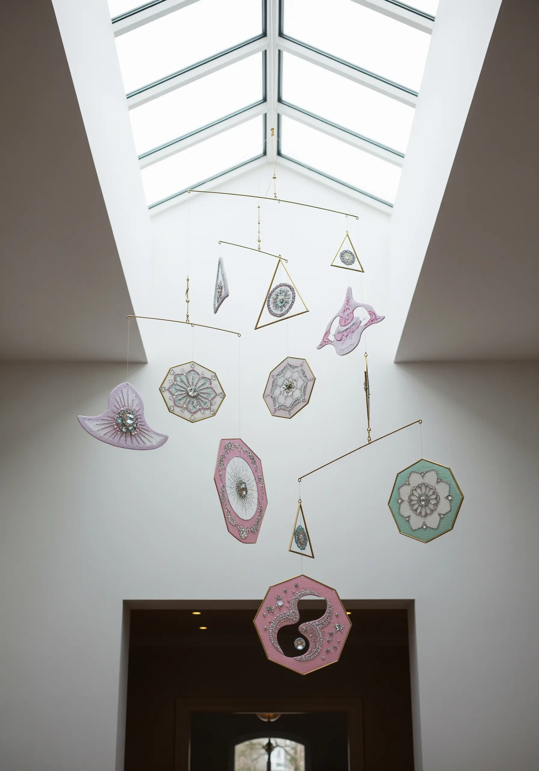 A delicate mobile hanging from a skylight, with embroidered and beaded shapes on sheer, pink-tinted fabric.