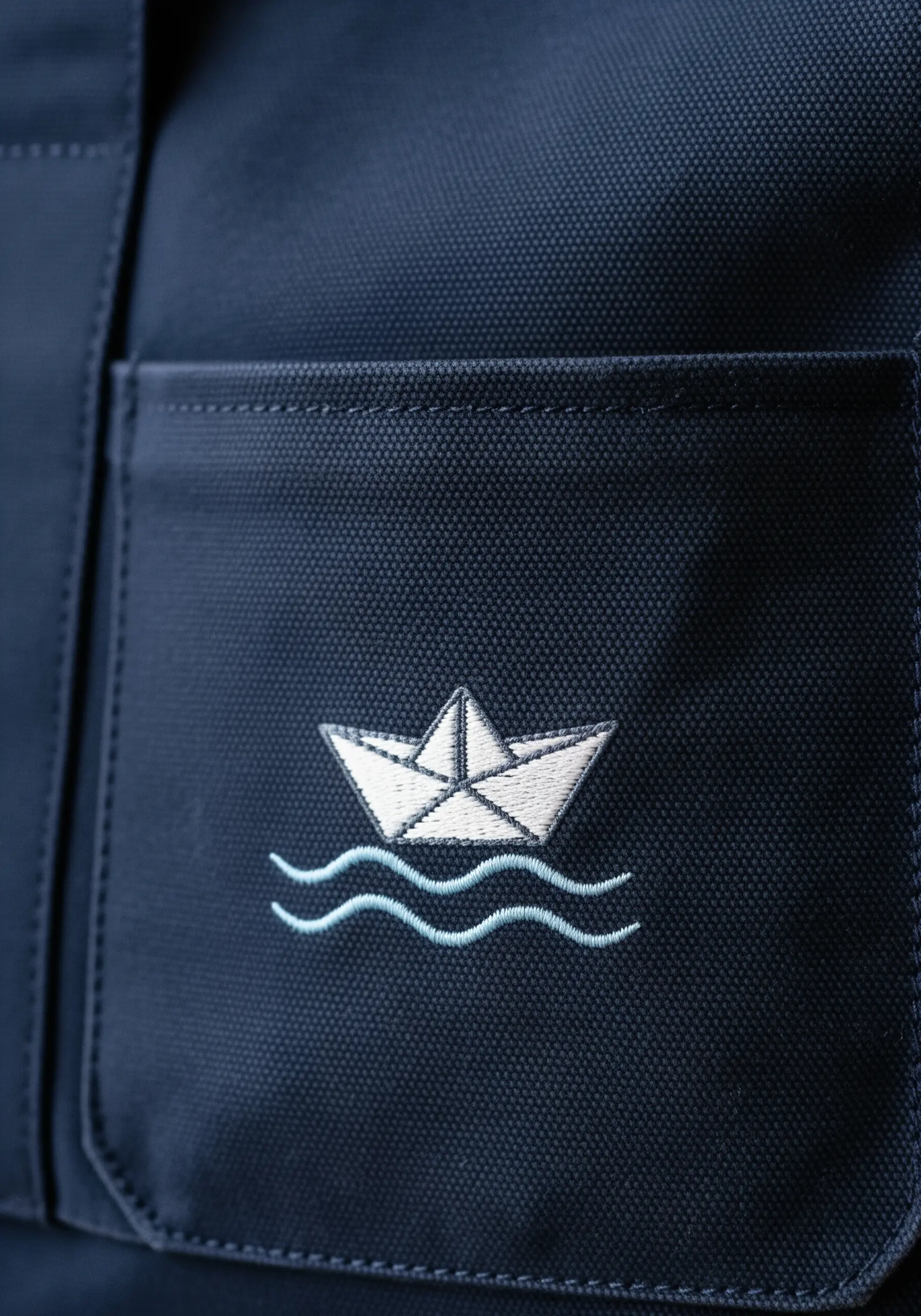 A white line-art embroidered paper boat on the pocket of a dark navy blue tote bag.