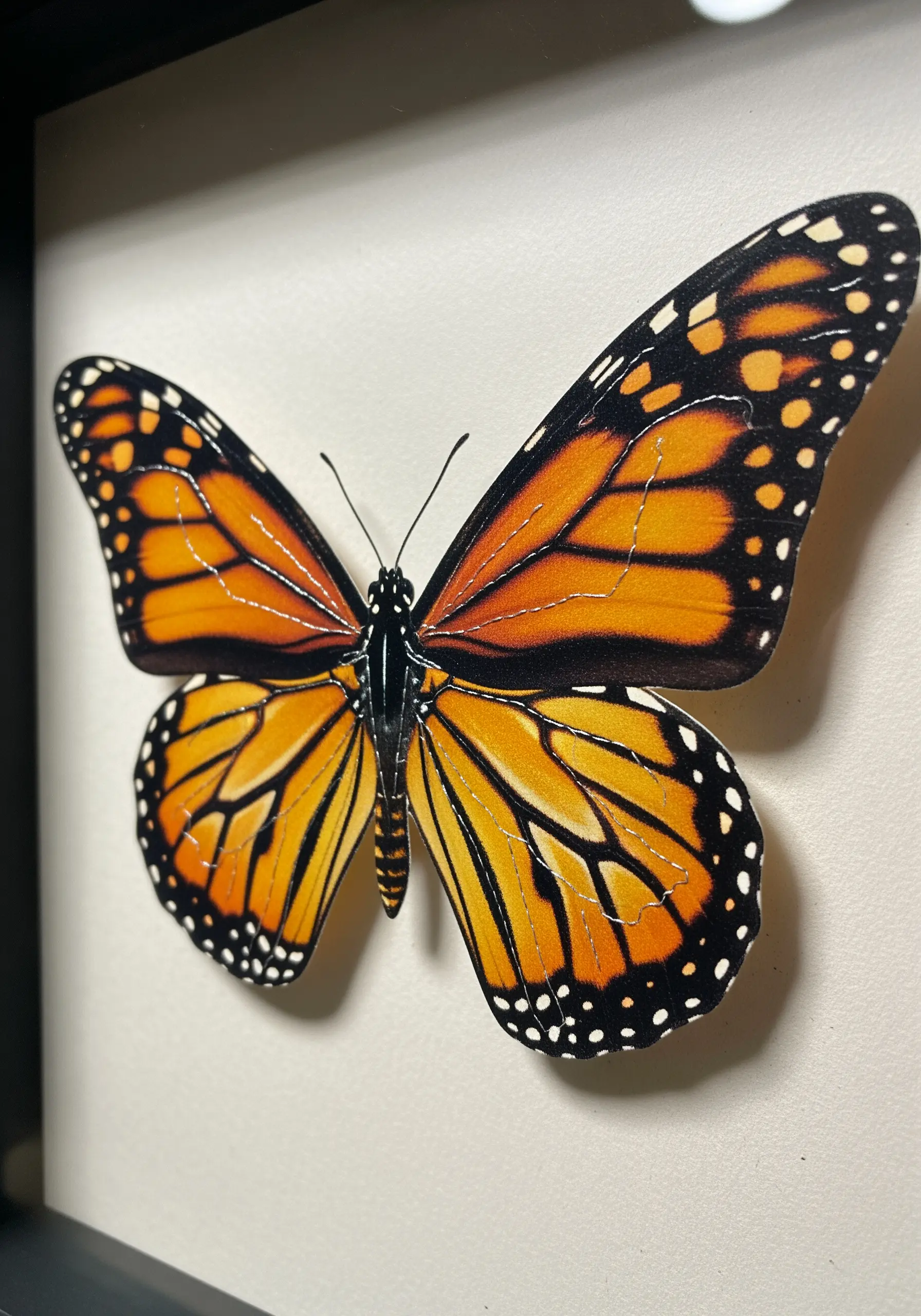 A realistic paper monarch butterfly with fine black stitching along its wing veins, mounted in a frame.