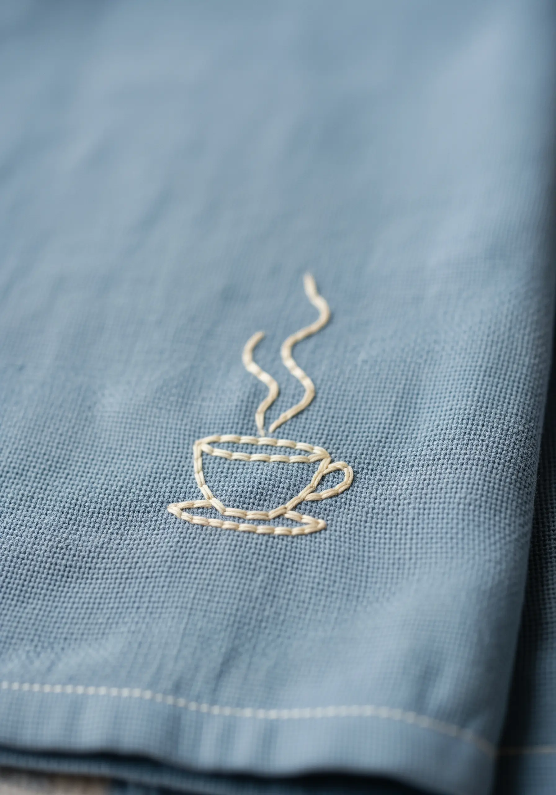 A minimalist outline of a steaming coffee cup stitched in white on a blue linen towel.