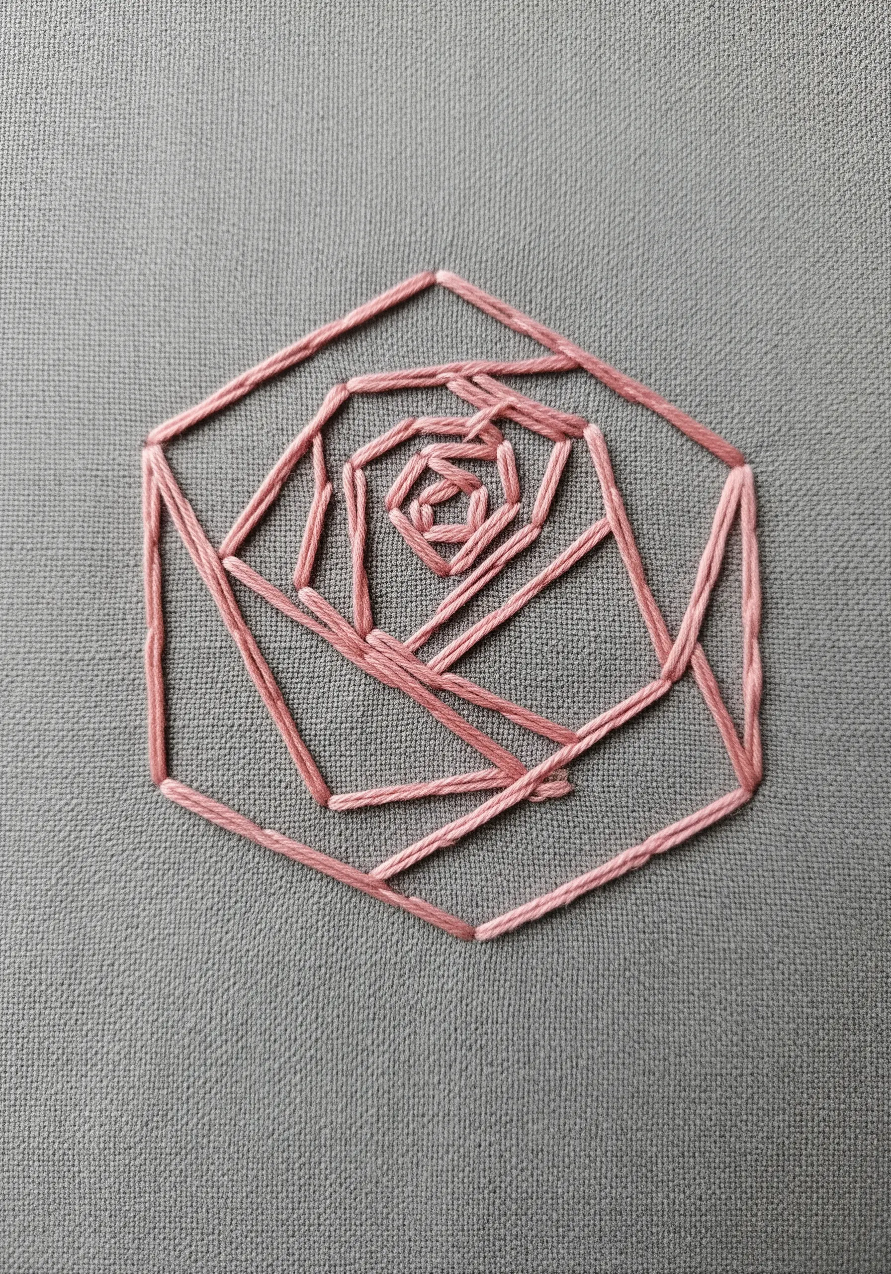 A modern, geometric rose embroidered with pink thread on grey fabric.