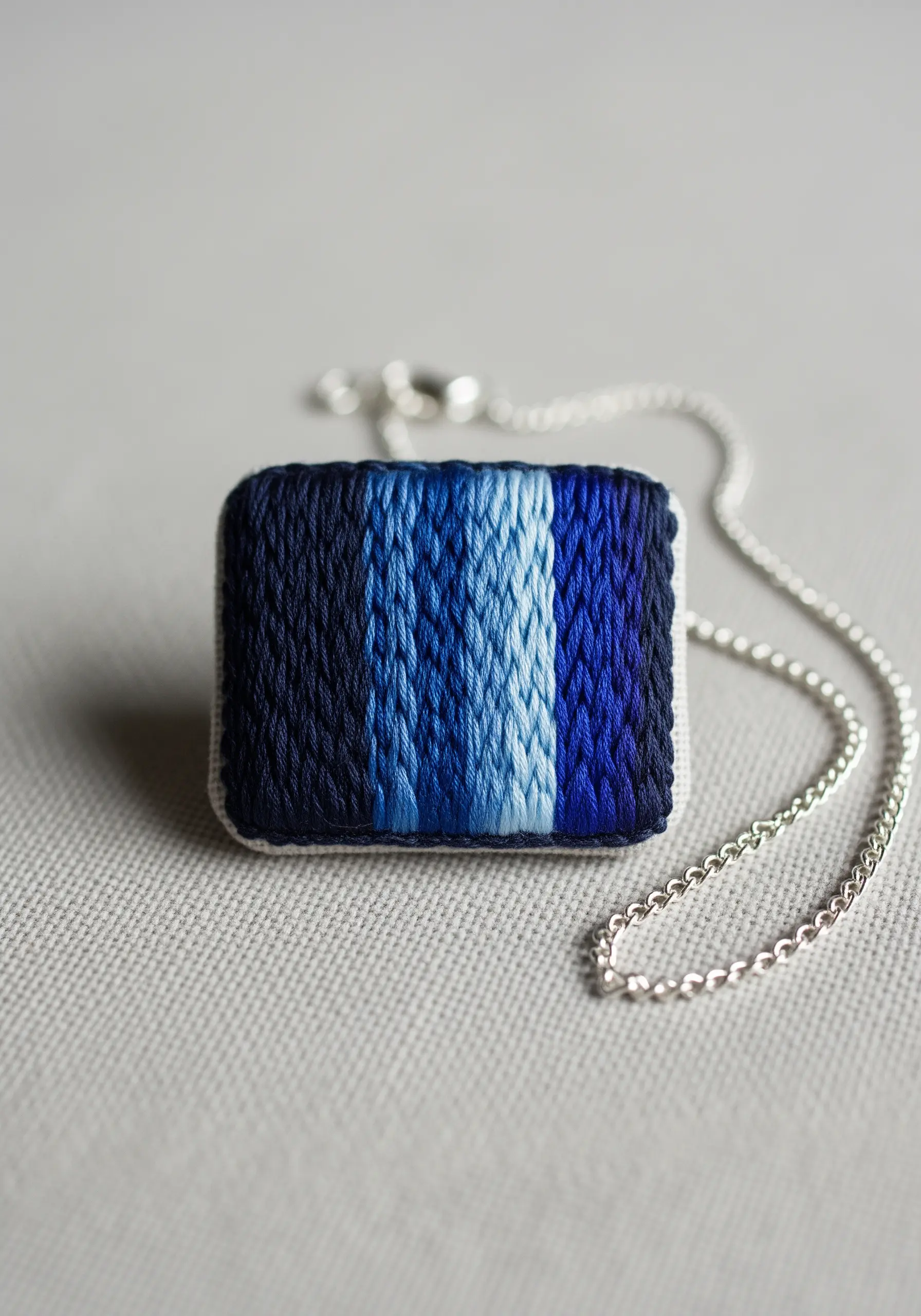 A square pendant with vertical stripes of thread painting in shades of blue.