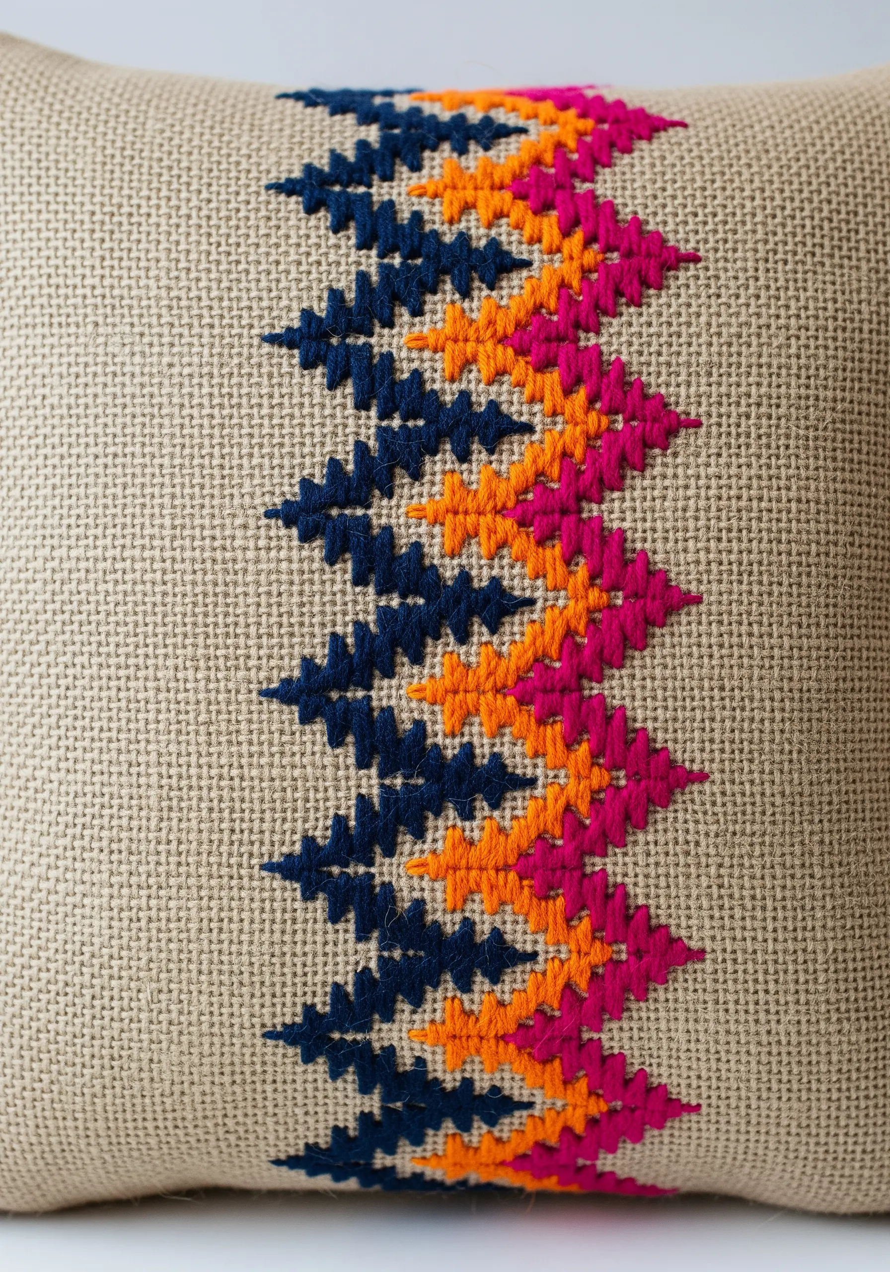 Geometric zig-zag pattern in cross stitch using pink, orange, and navy on burlap.