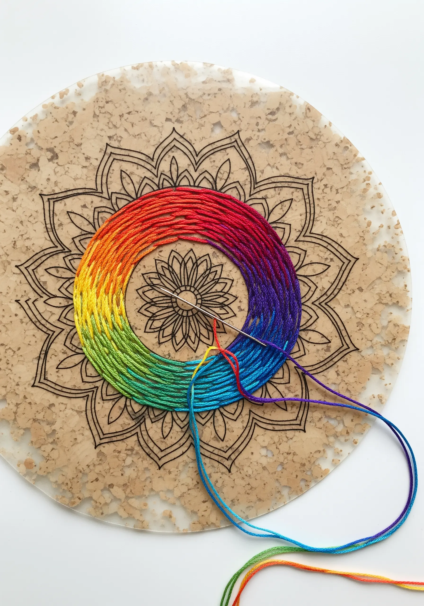 Rainbow-colored thread being wrapped around a circular cork board with a laser-etched mandala design.