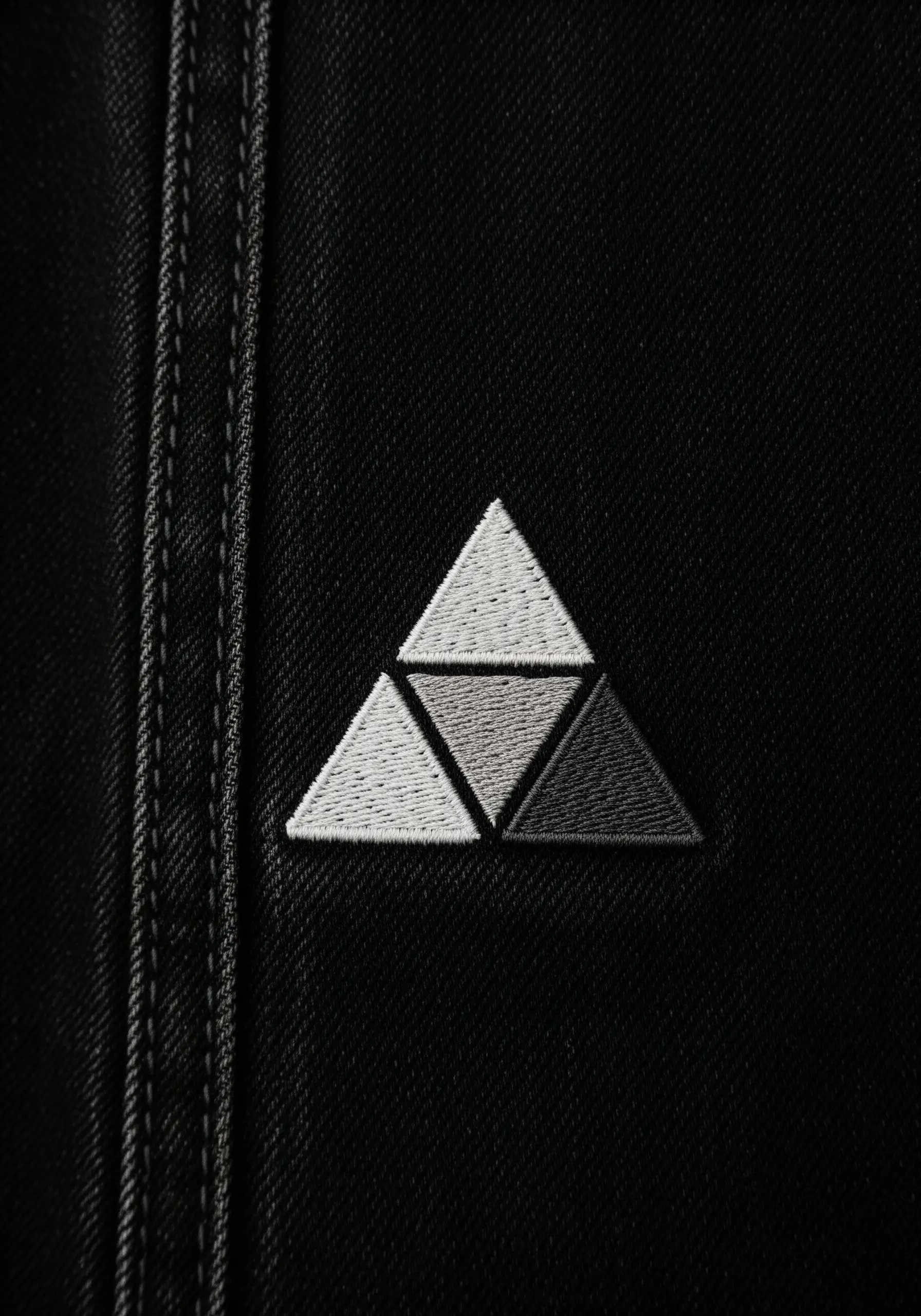 A geometric motif of triangles filled with a grayscale gradient of thread on black denim.