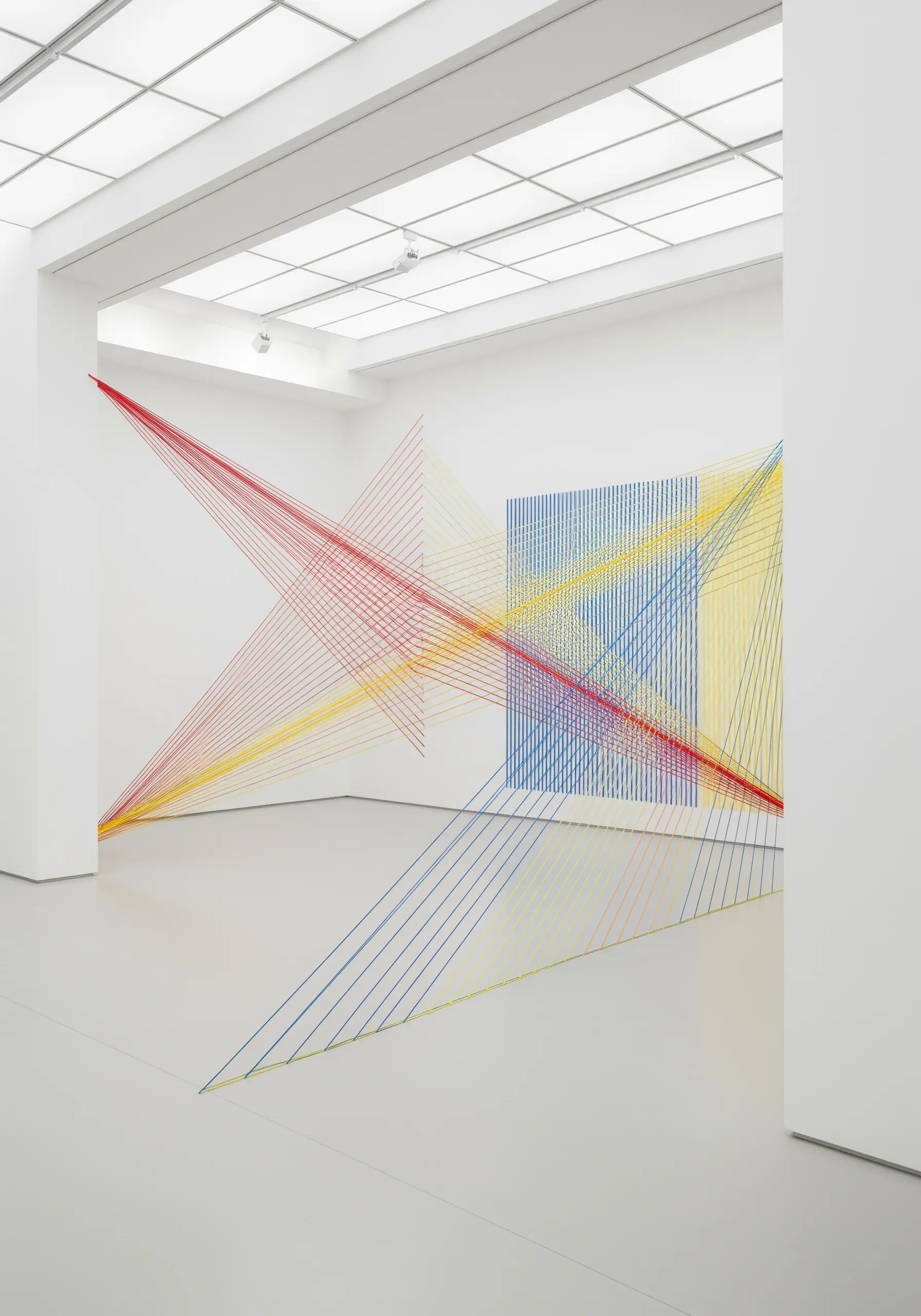 Minimalist fiber art of red, yellow, and blue threads stretched across a white gallery.