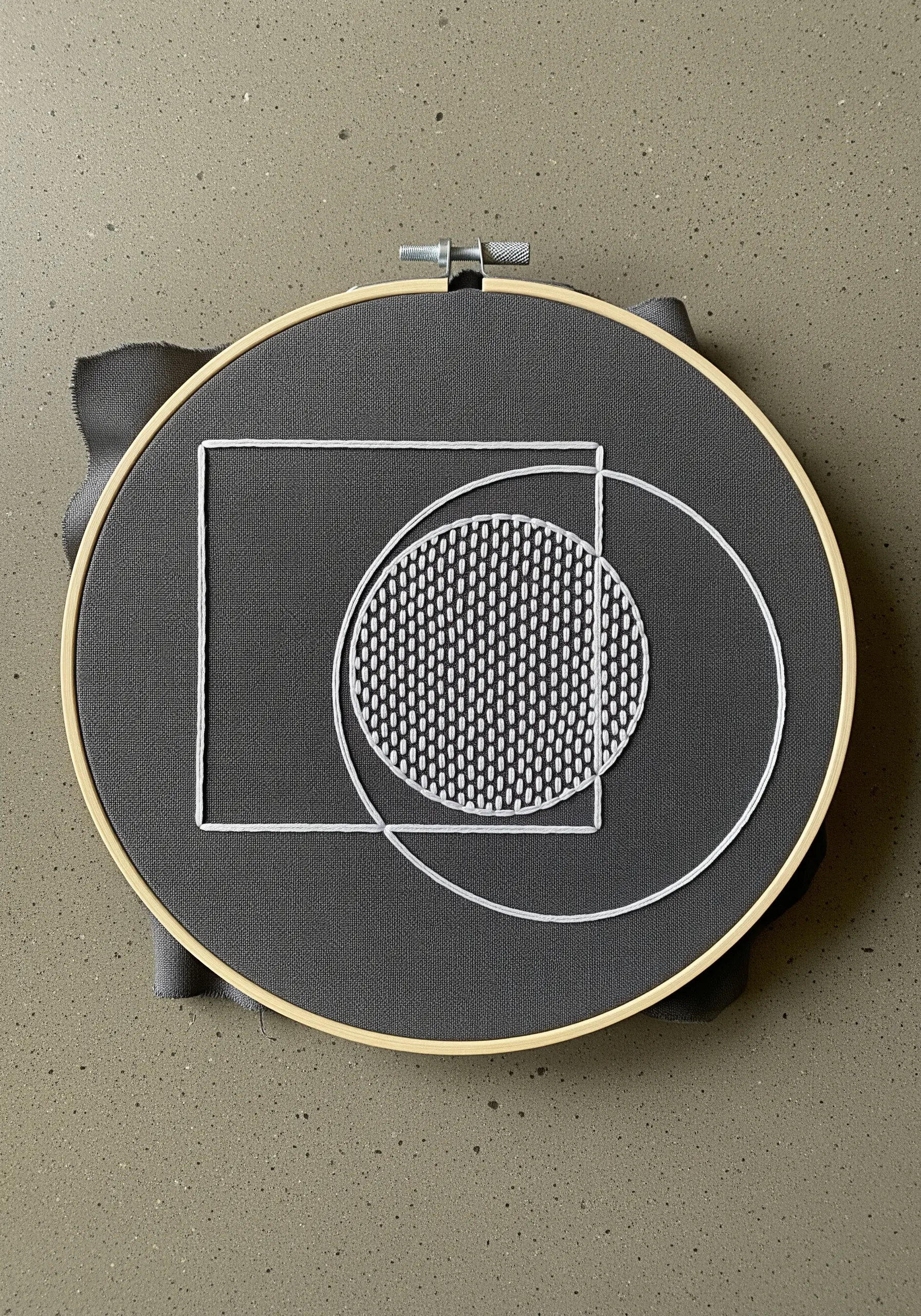 Abstract embroidery of a white square and circle with a textured fill on grey fabric.