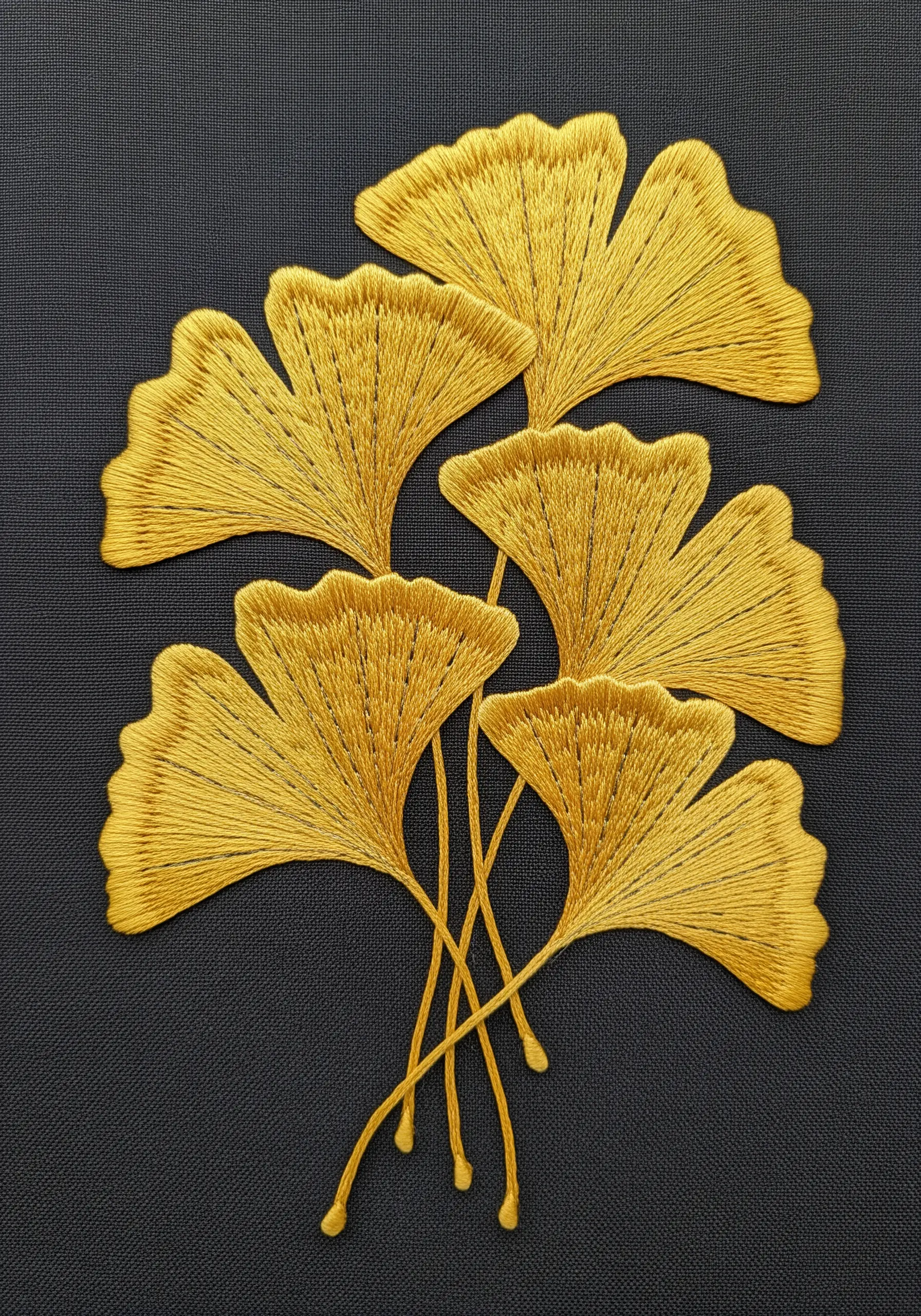 Golden yellow satin-stitched ginkgo leaves on a dark grey fabric background.
