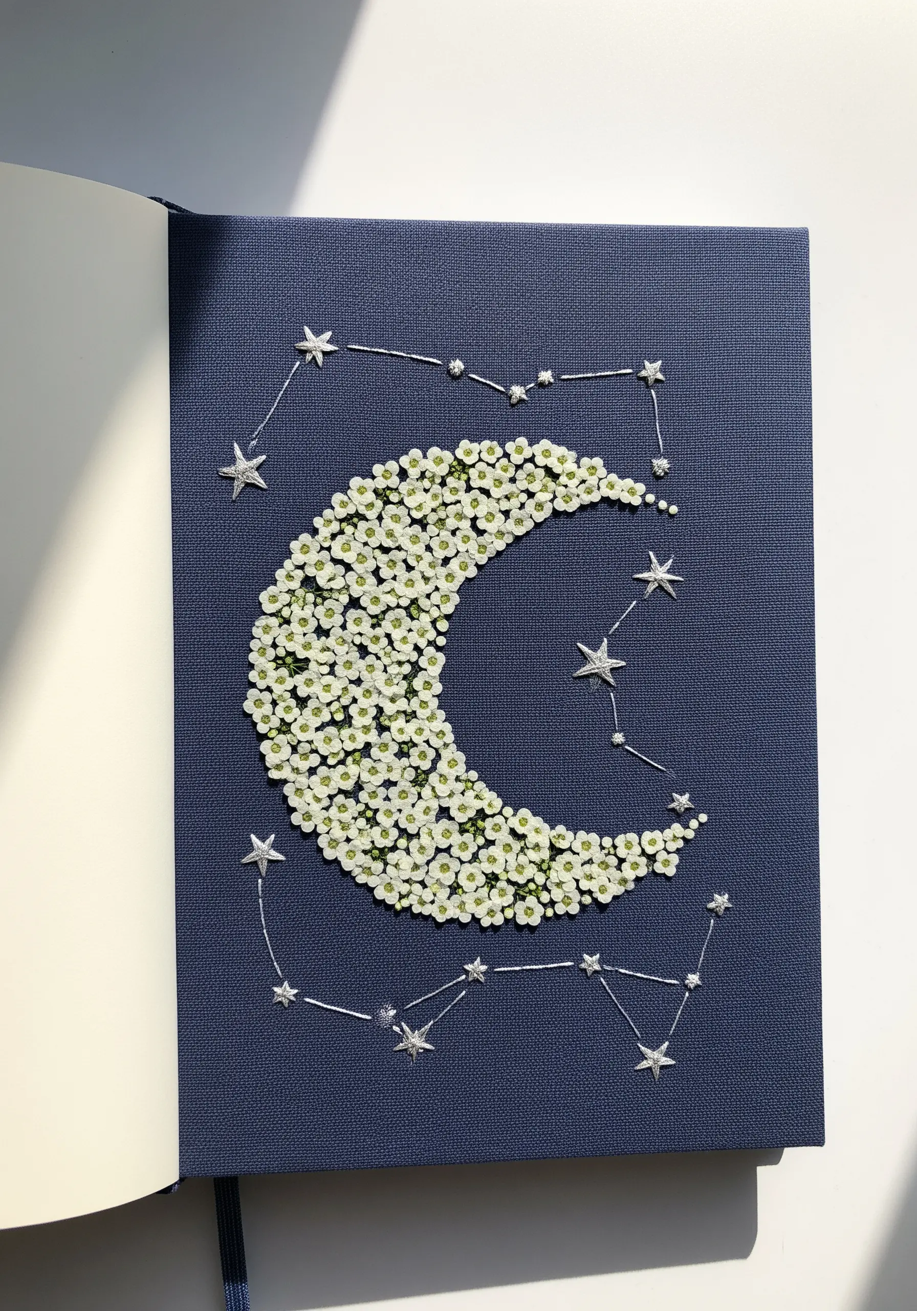 A crescent moon on a blue journal, outlined in silver thread and filled with tiny white flowers.