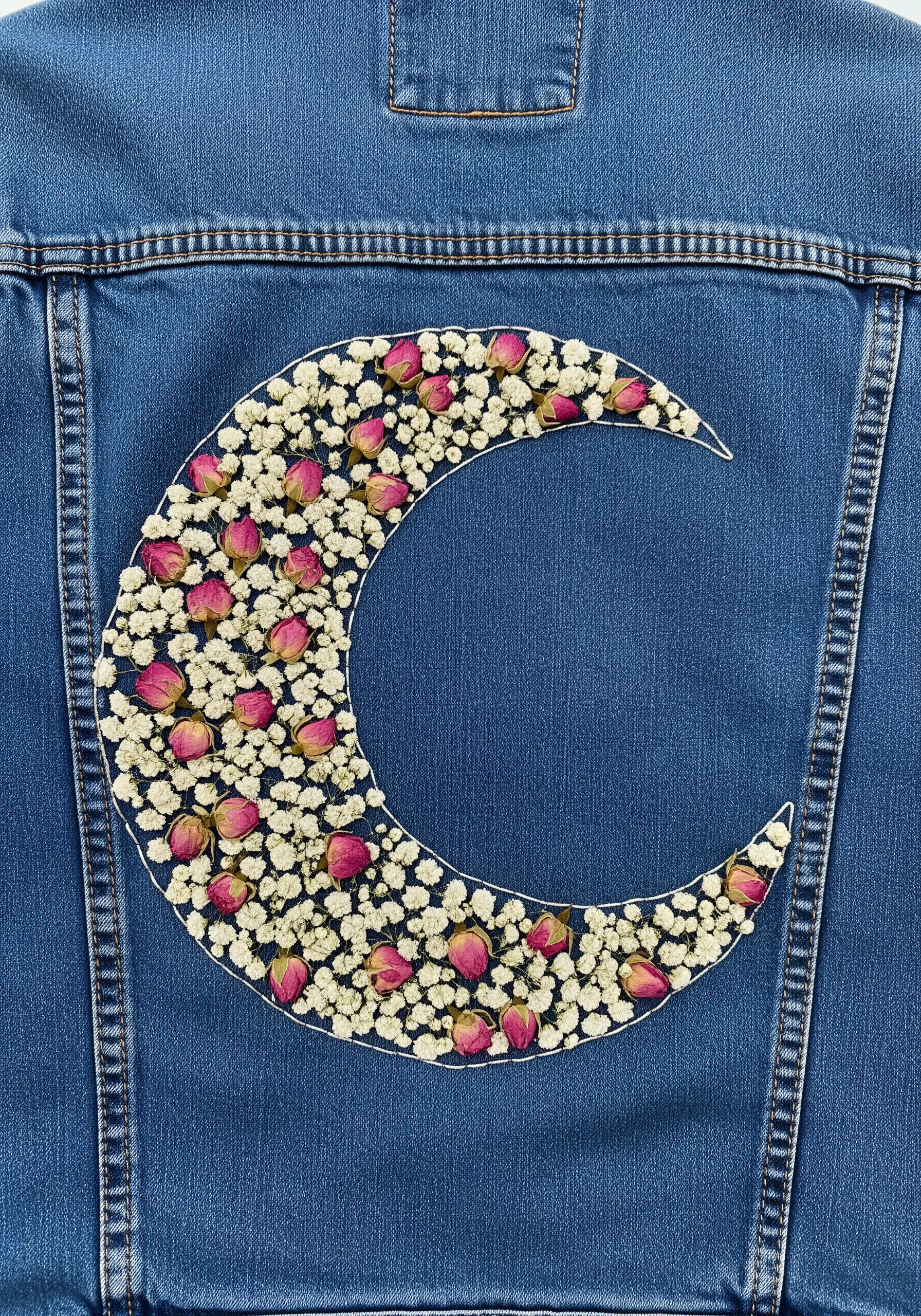A crescent moon on a denim jacket, outlined in white thread and filled with tiny rosebuds.