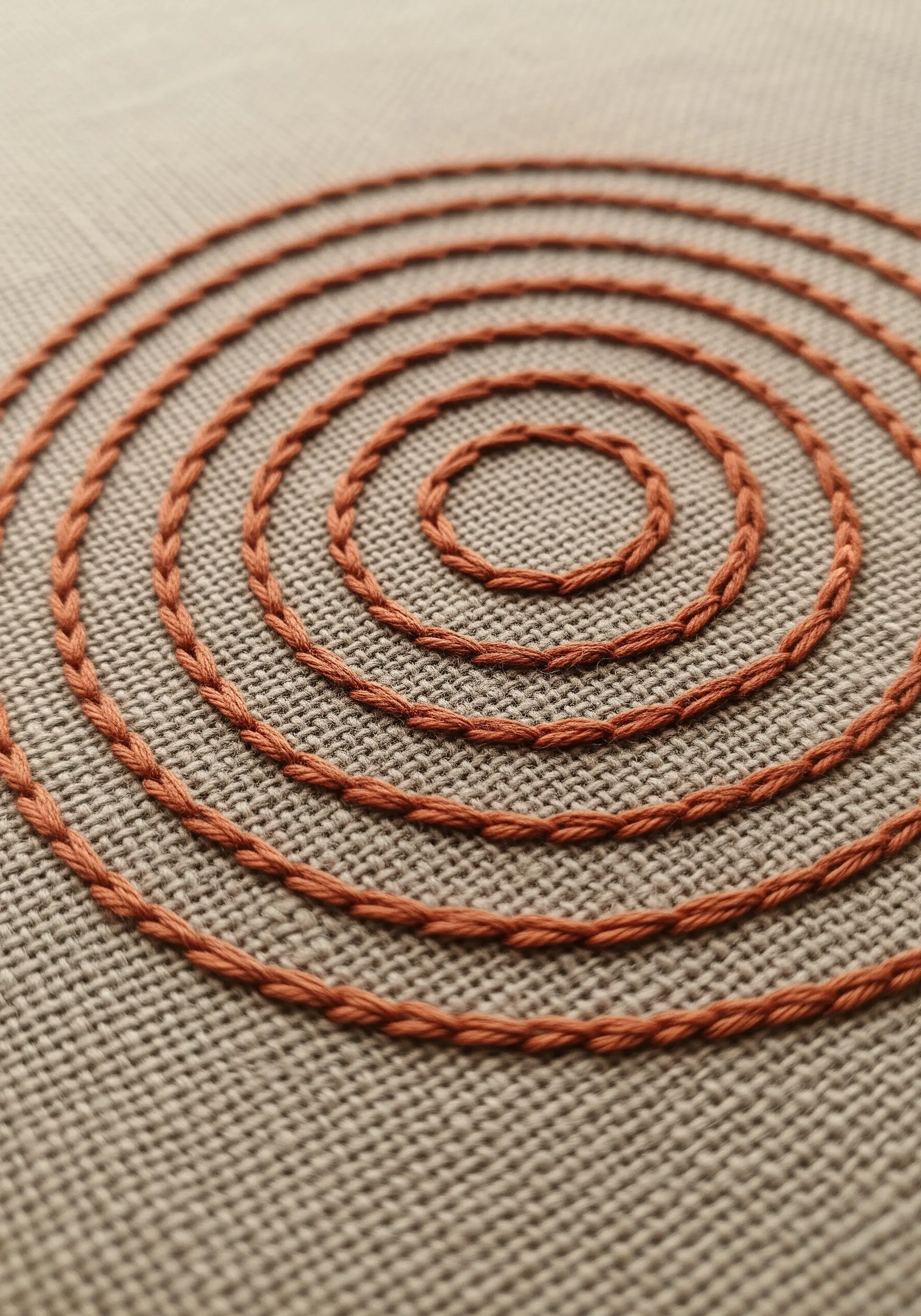 A clean spiral of terracotta-colored chain stitch on natural linen fabric.