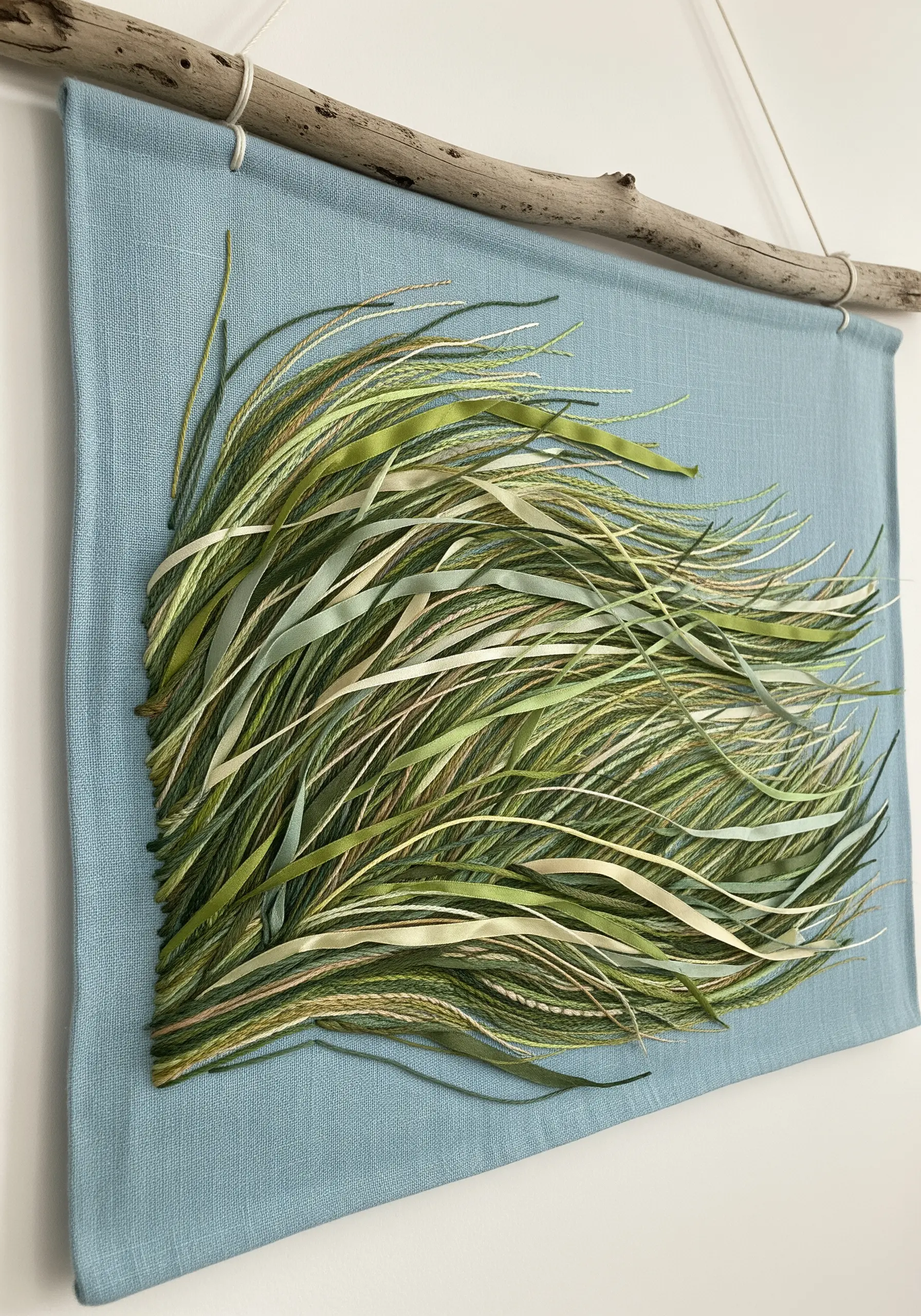 Wall hanging with long, loose green and beige threads cascading to mimic flowing grass.