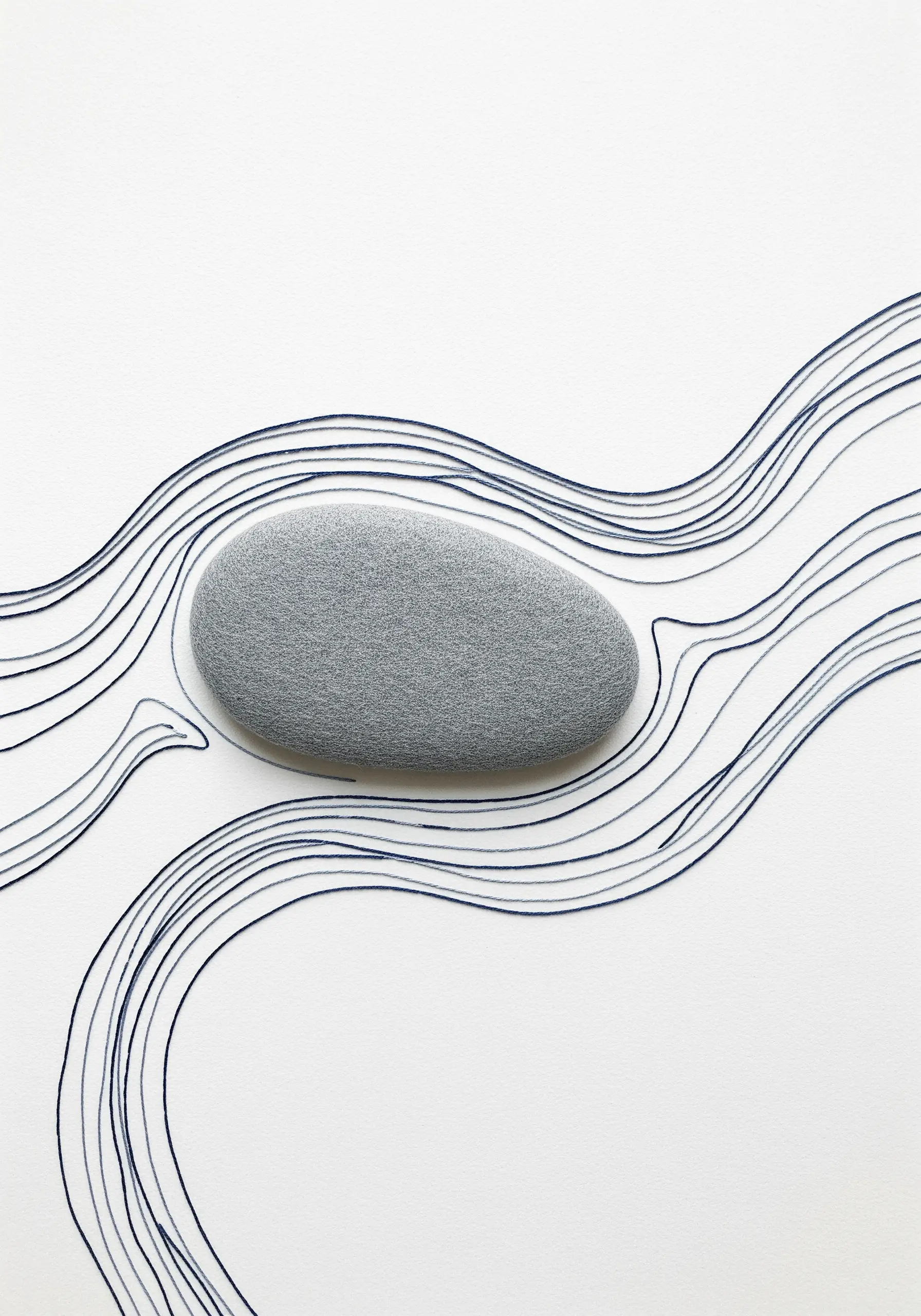 Minimalist embroidery showing fine blue lines flowing around a smooth gray stone on white fabric.