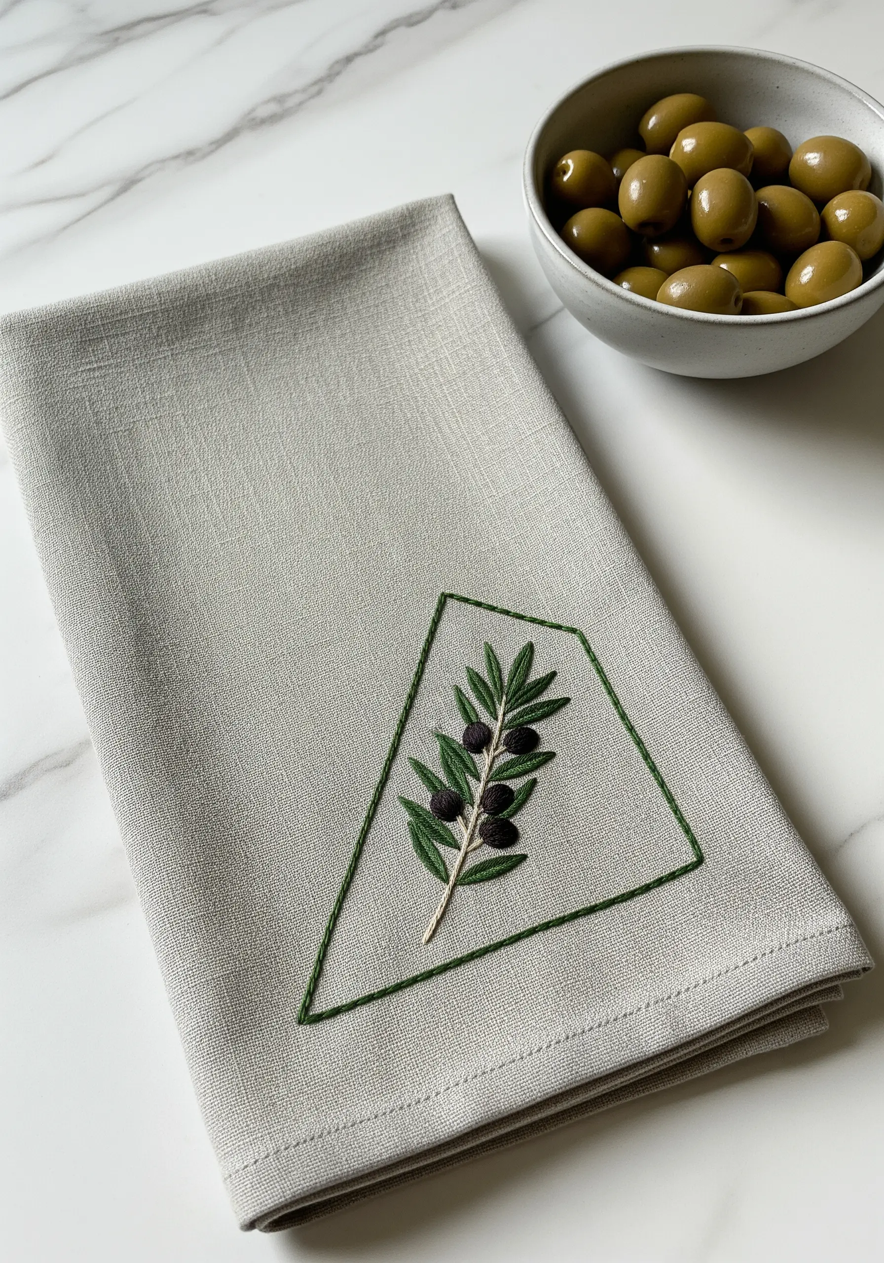 A single olive branch embroidered inside a geometric green border on a grey napkin.