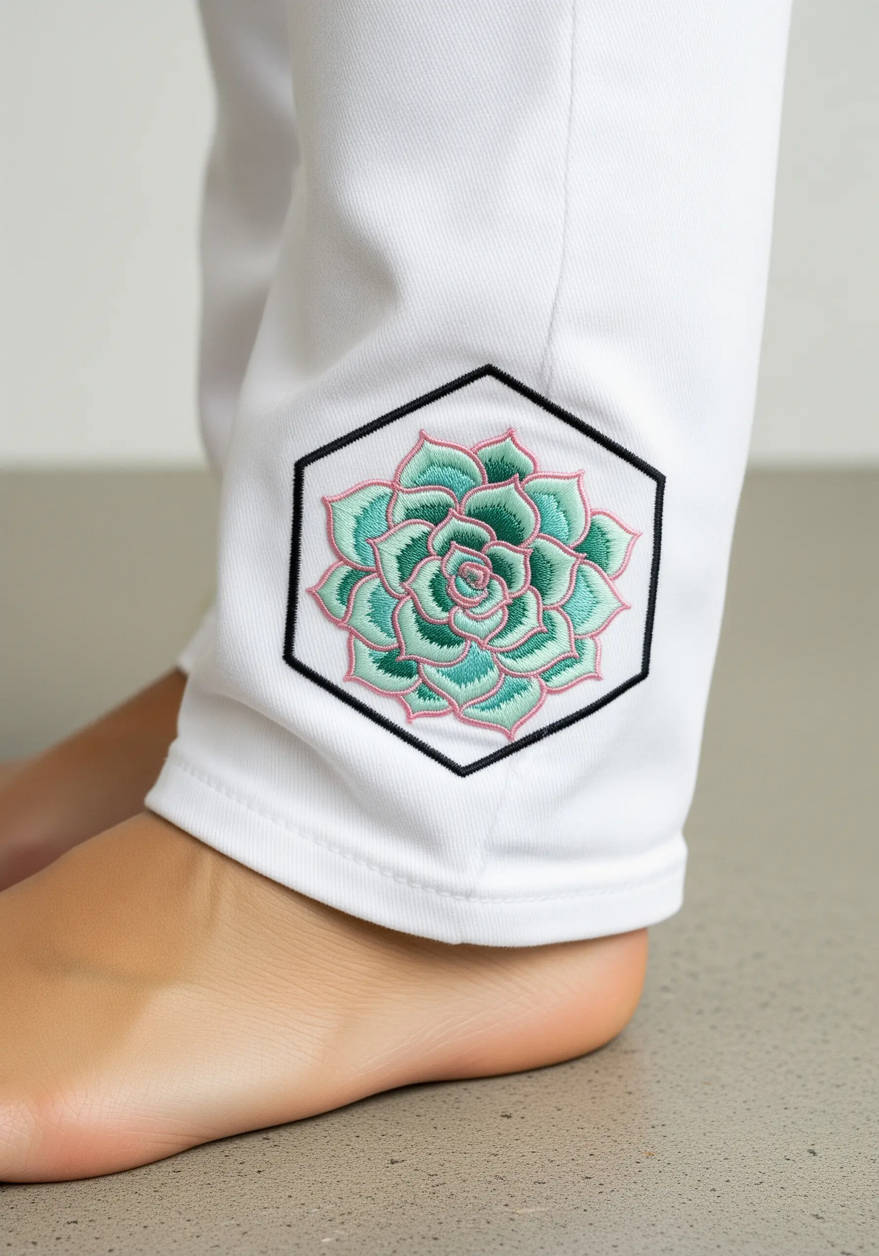 An embroidered succulent with pink and green gradients, framed in a black hexagon on white pants.