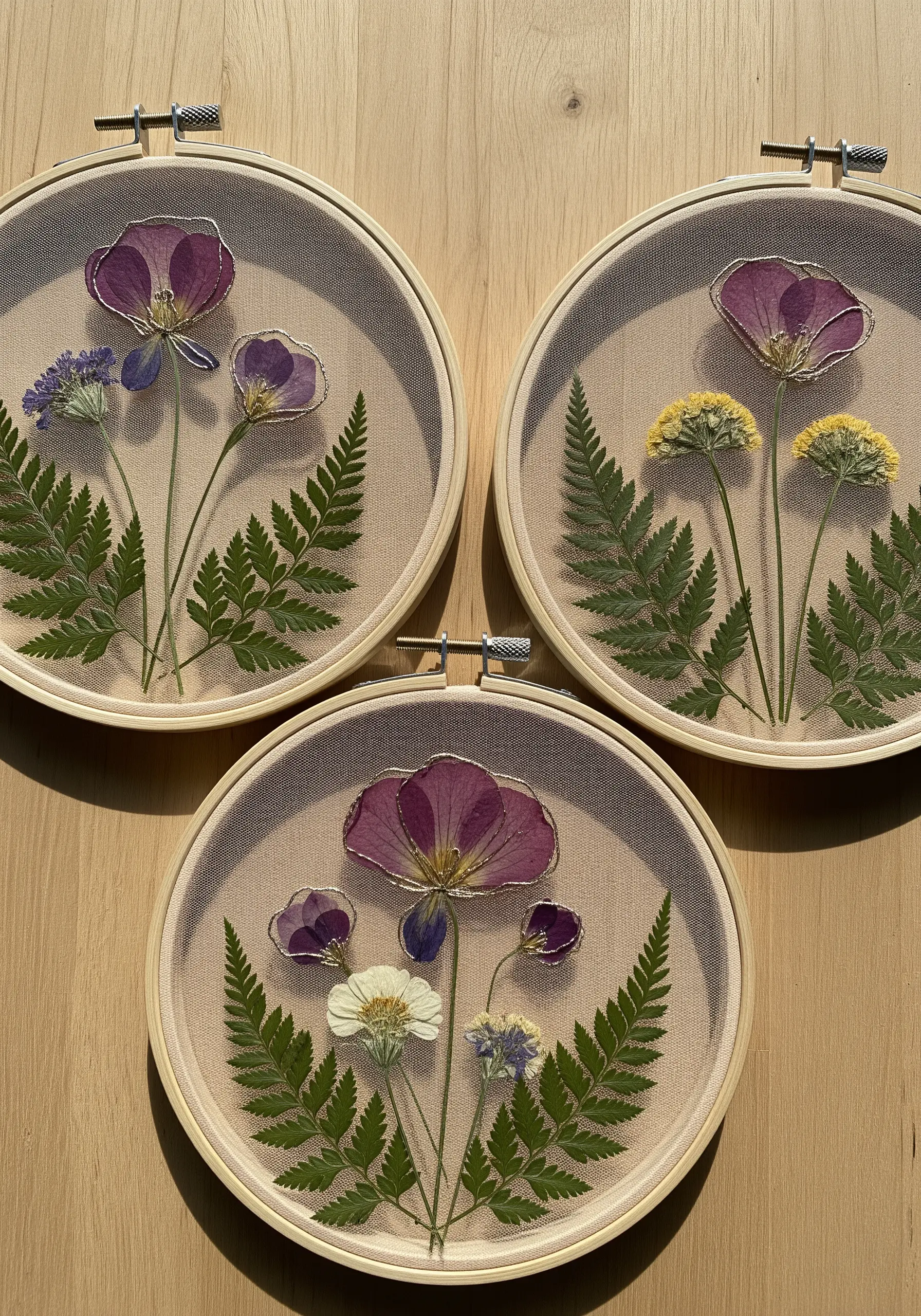 Pressed purple flowers and green ferns in embroidery hoops, outlined with fine gold metallic thread.