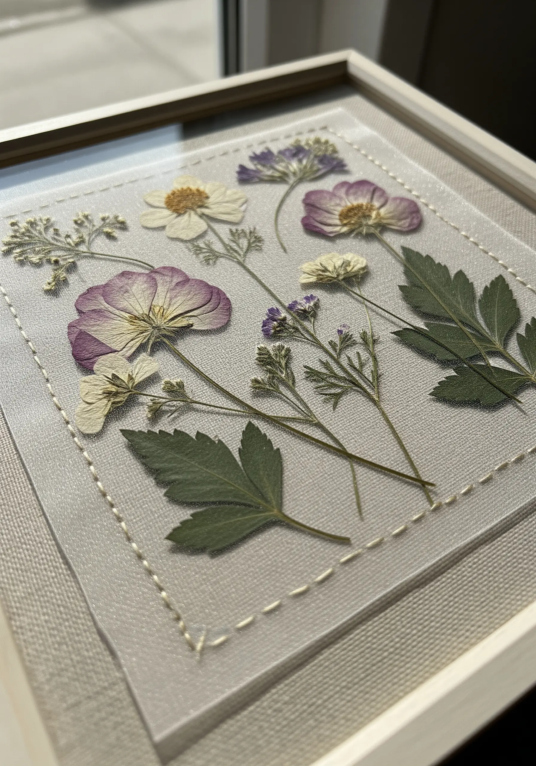 Pressed flowers secured onto sheer fabric with a simple running stitch border inside a frame.