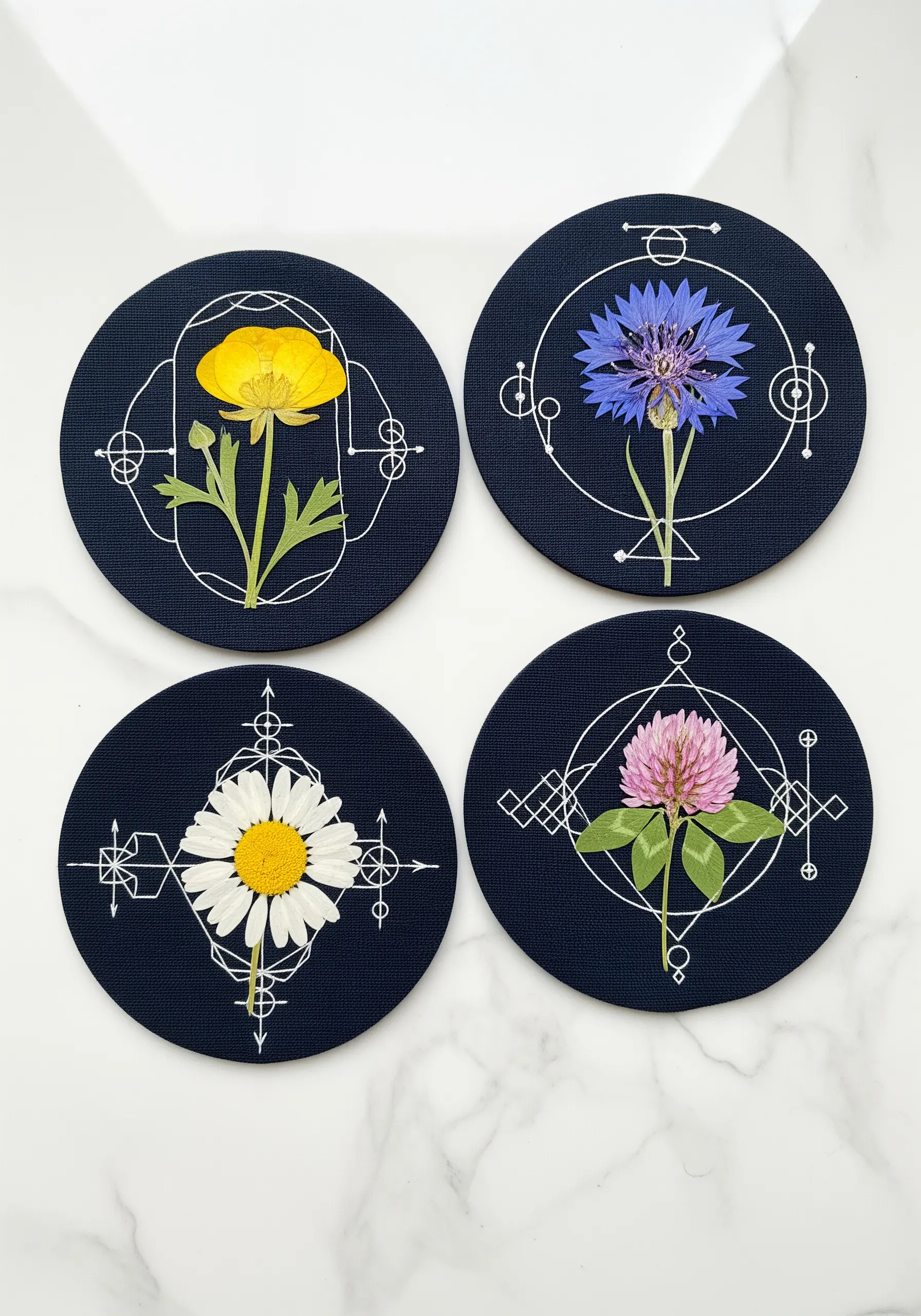 Four coasters, each with a pressed flower framed by white geometric line embroidery.