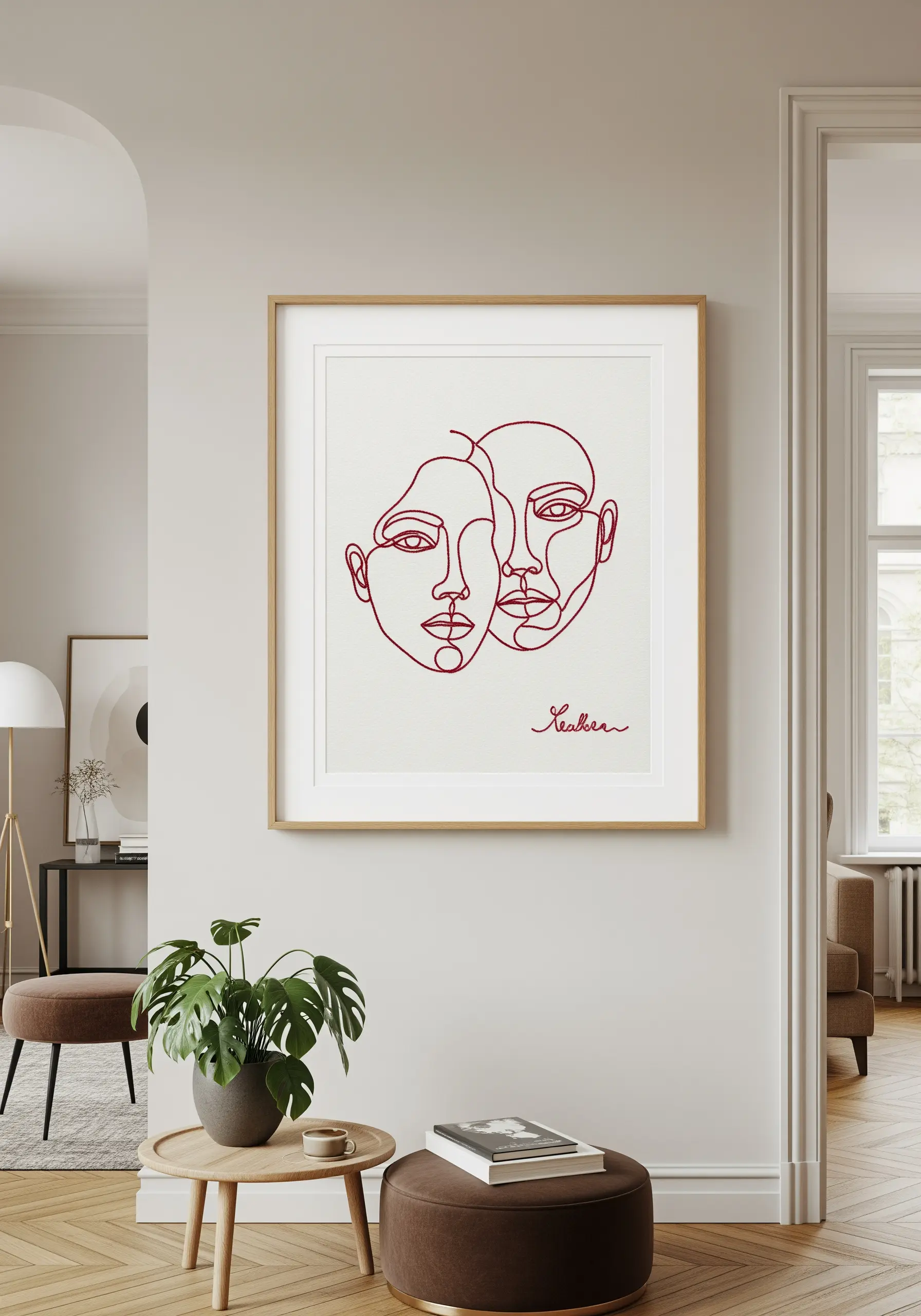 A simple red line-art embroidery of two faces, framed with a large white mat in a modern room.