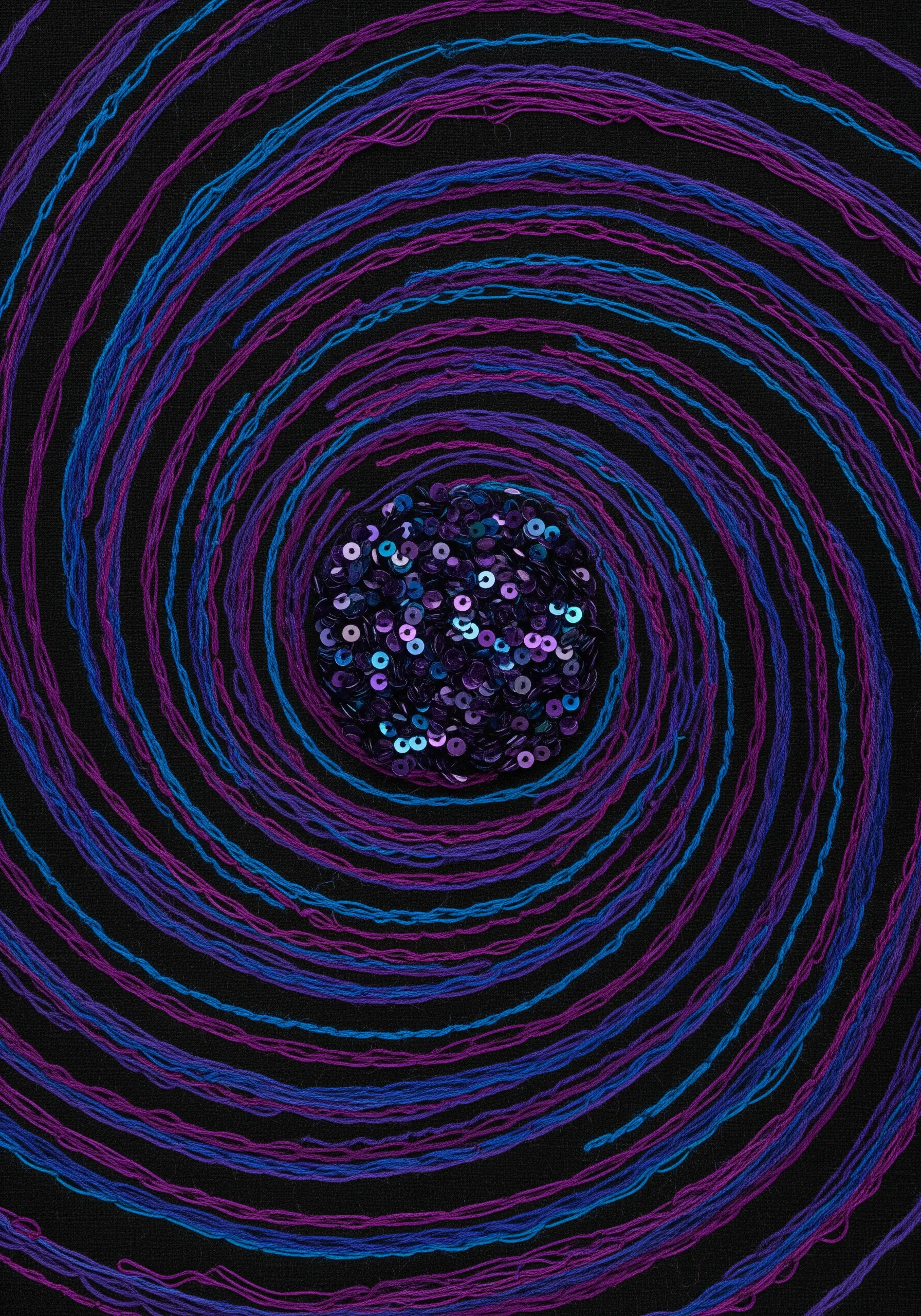 A spiral of purple and blue threads on black fabric, with a central cluster of sequins.