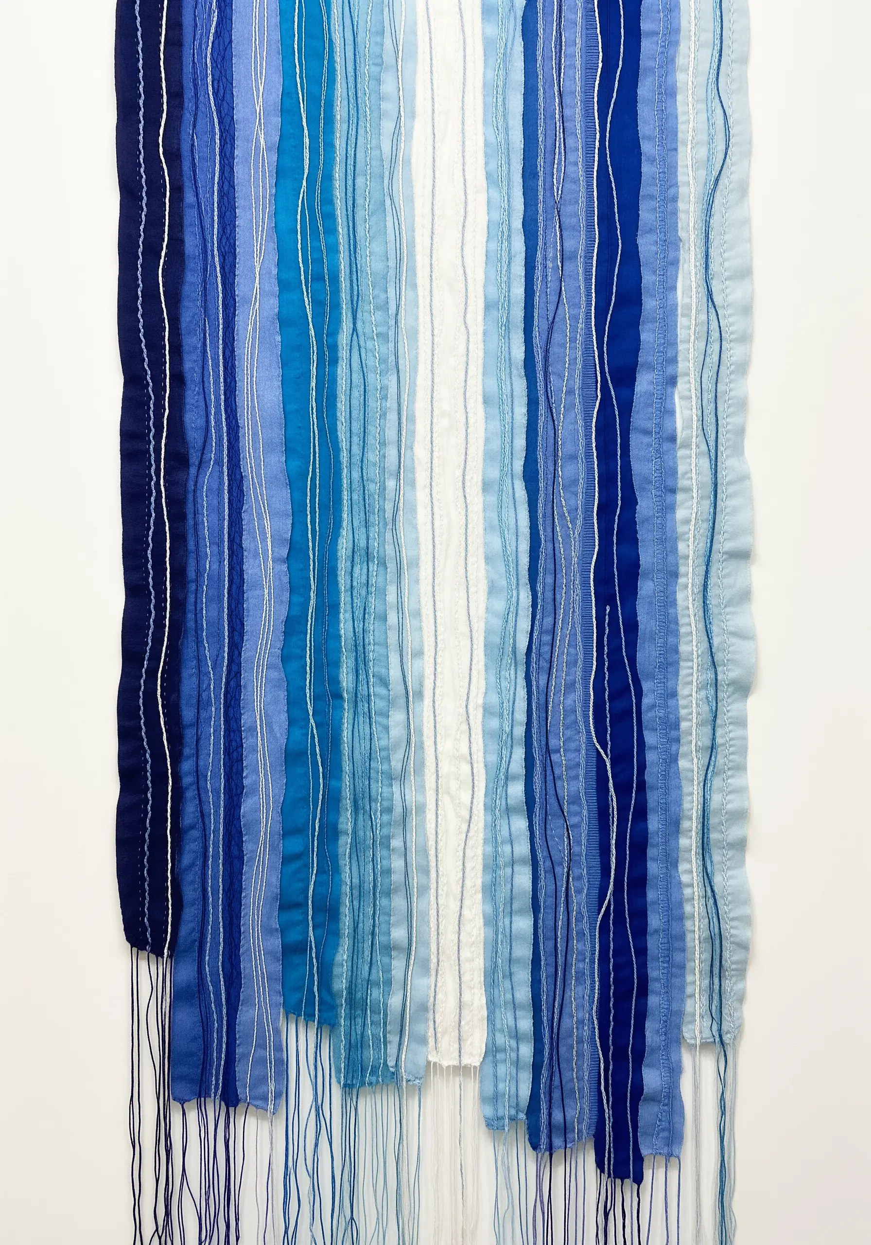 A wall hanging made of vertical blue fabric strips arranged in a gradient with loose threads.