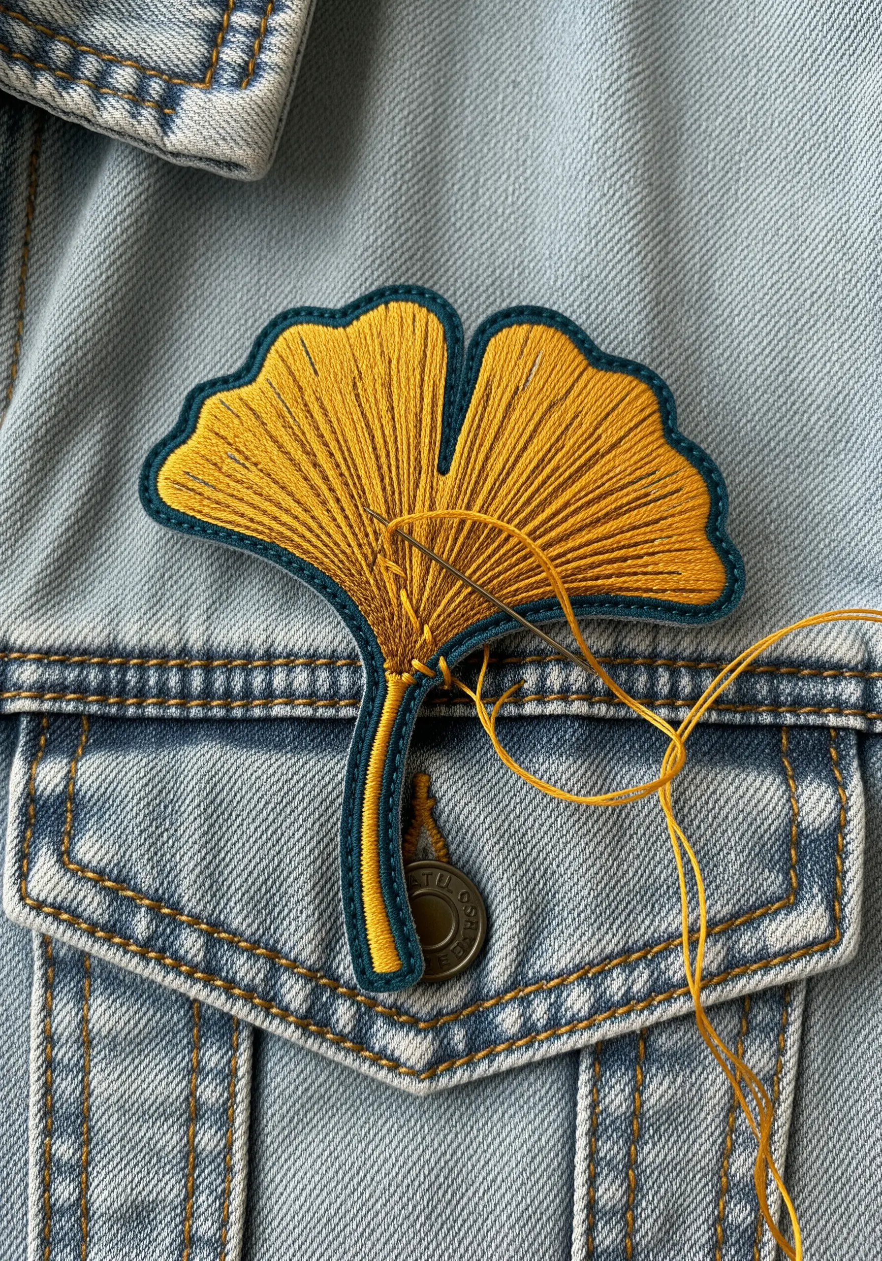 A vibrant yellow ginkgo leaf patch being stitched onto a denim jacket.