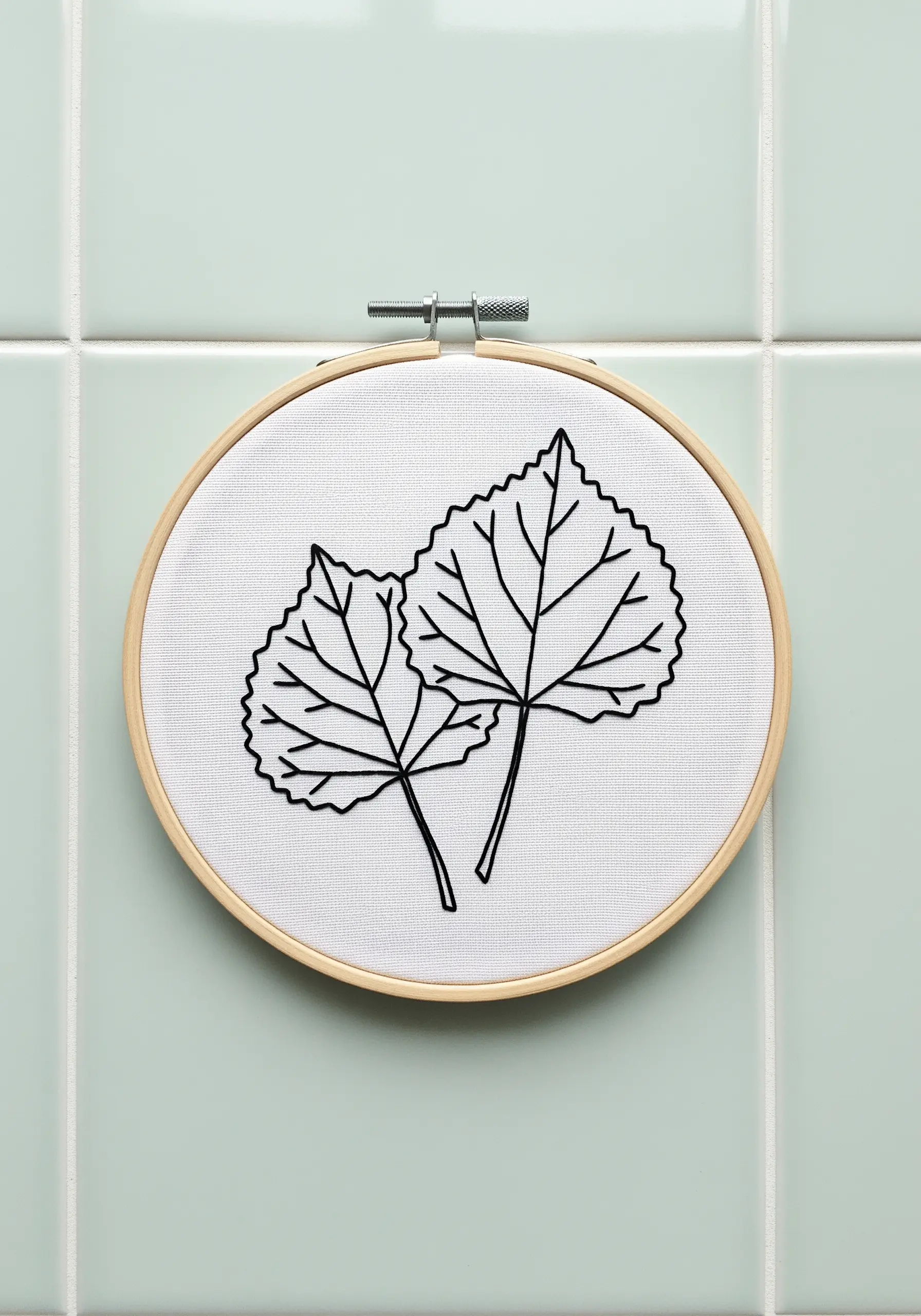 Two leaves embroidered in a bold black outline on crisp white fabric against a tiled wall.
