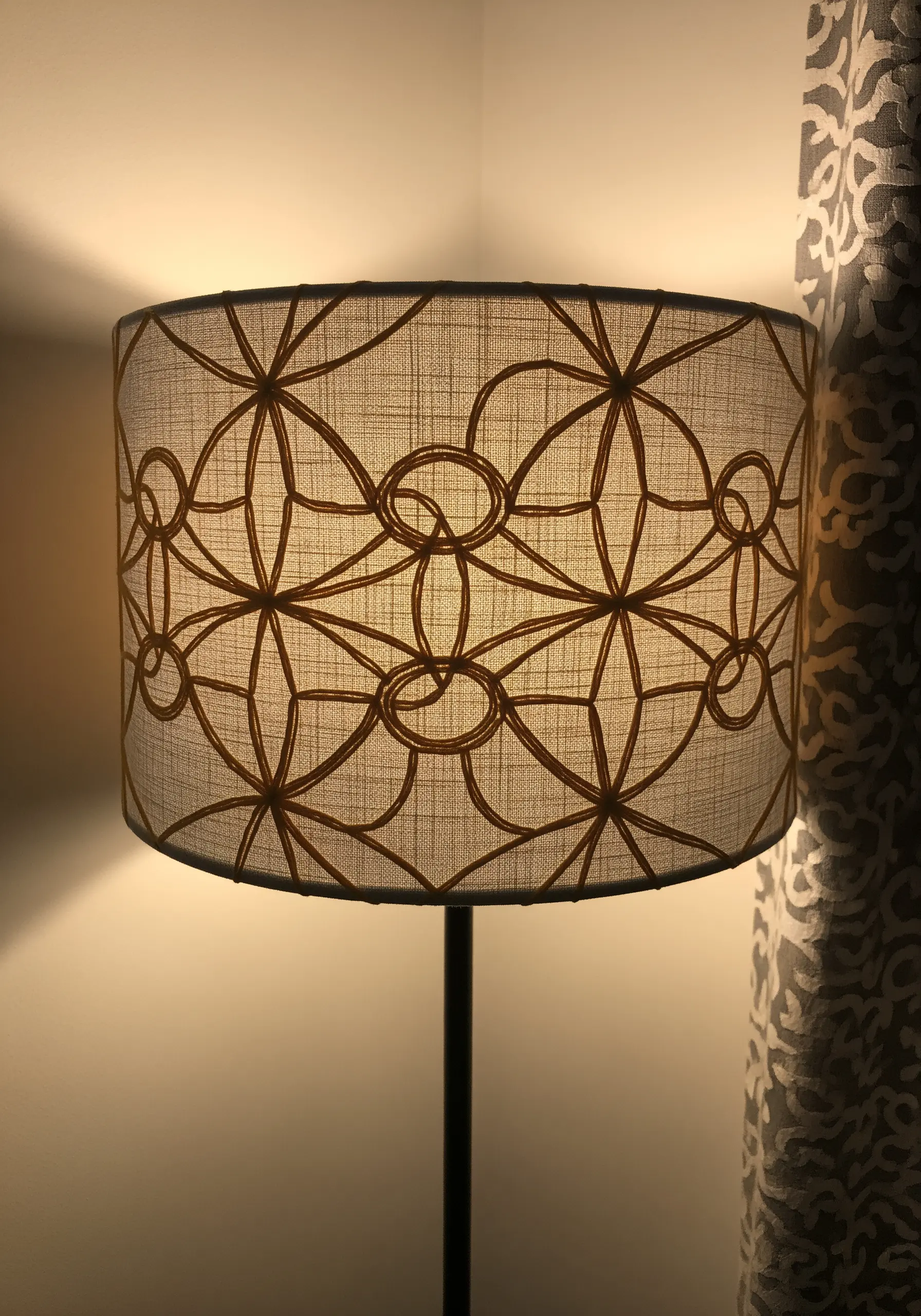 A lit lampshade with a geometric pattern embroidered in a golden-brown thread.
