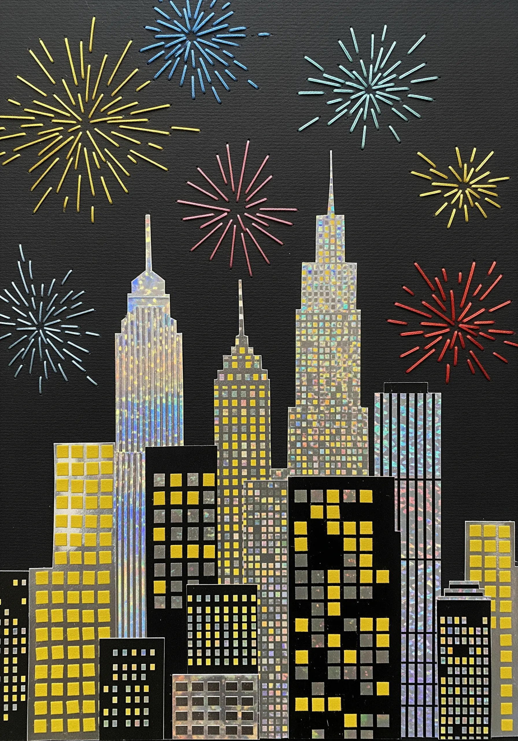 A paper collage of a city skyline at night with colorful embroidered fireworks.