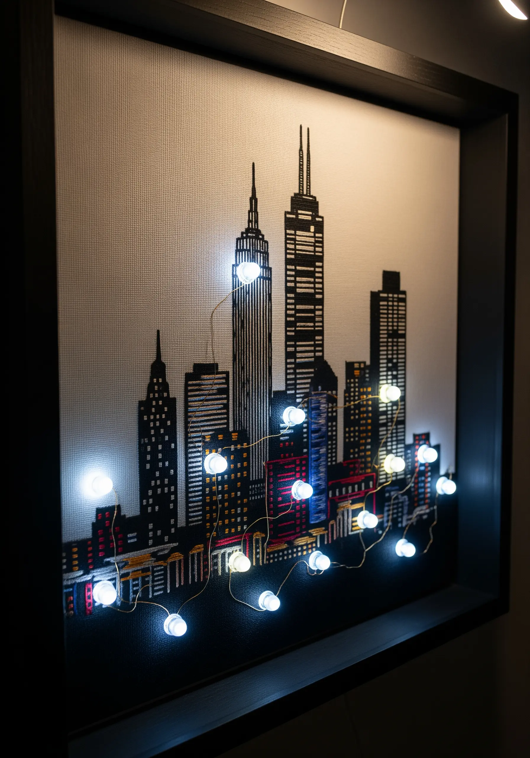 A framed cross-stitch of a city skyline with small white LED lights integrated into the design.