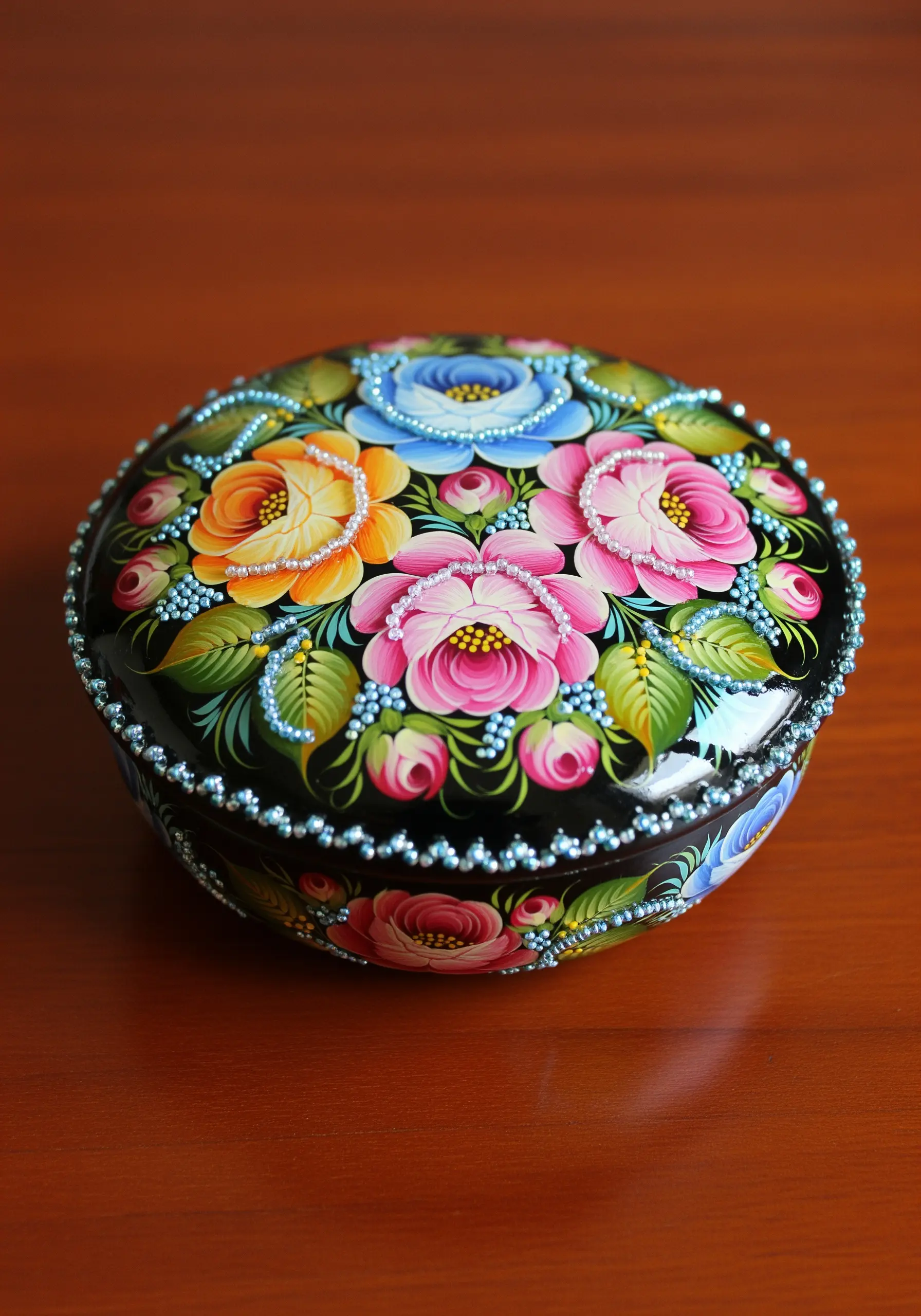 A round, black lacquered box painted with flowers and embellished with seed beads.