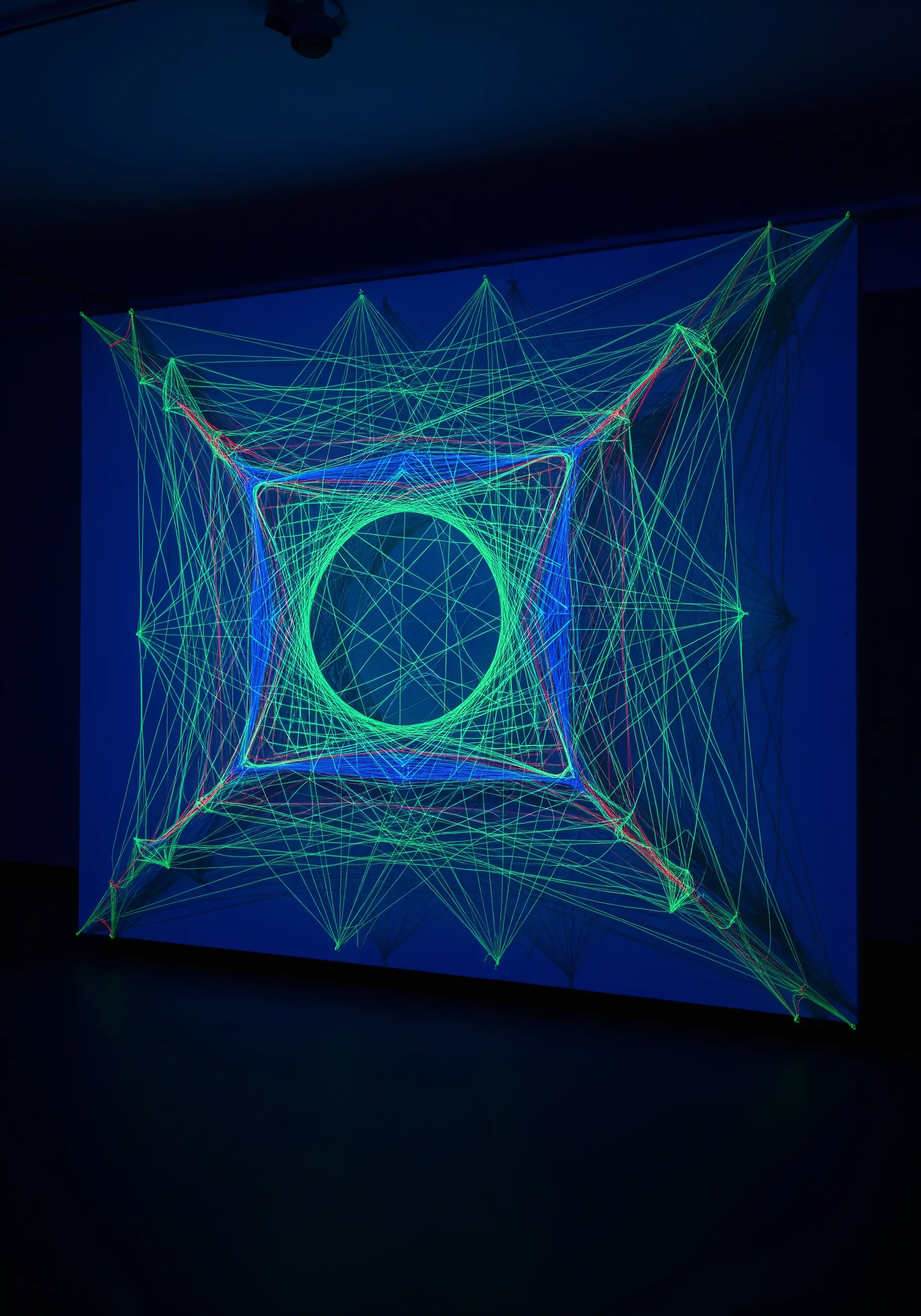 A large geometric string art installation glowing with bright green neon thread under a blacklight.
