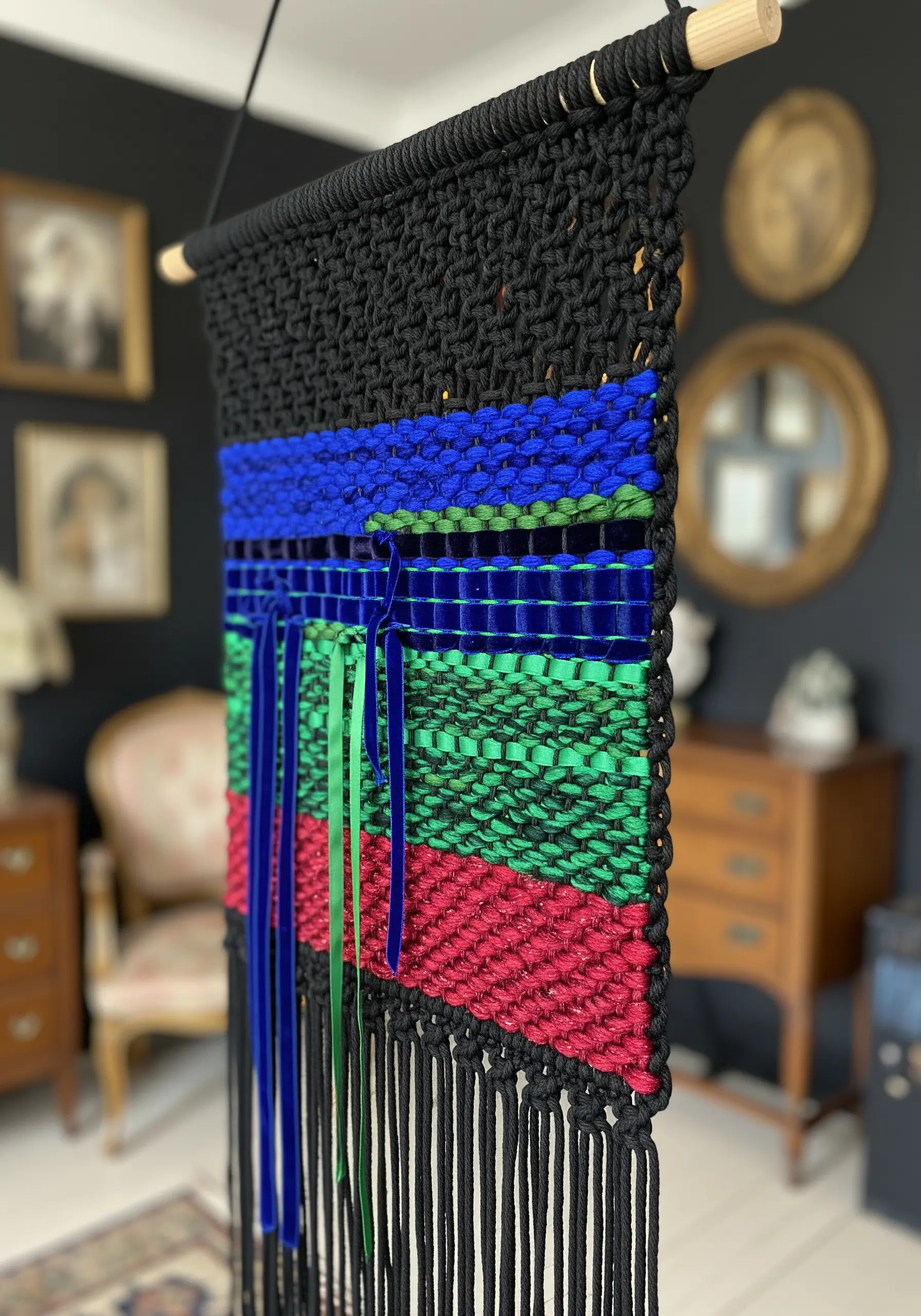 Black macrame wall hanging with color-blocked sections of woven blue, green, and red velvet ribbon.