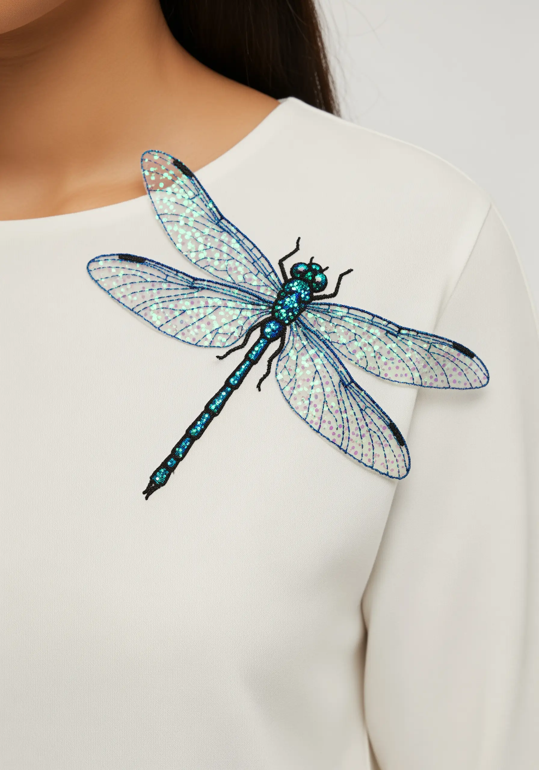 A large dragonfly with iridescent appliqué wings and a glitter thread body on a white top.