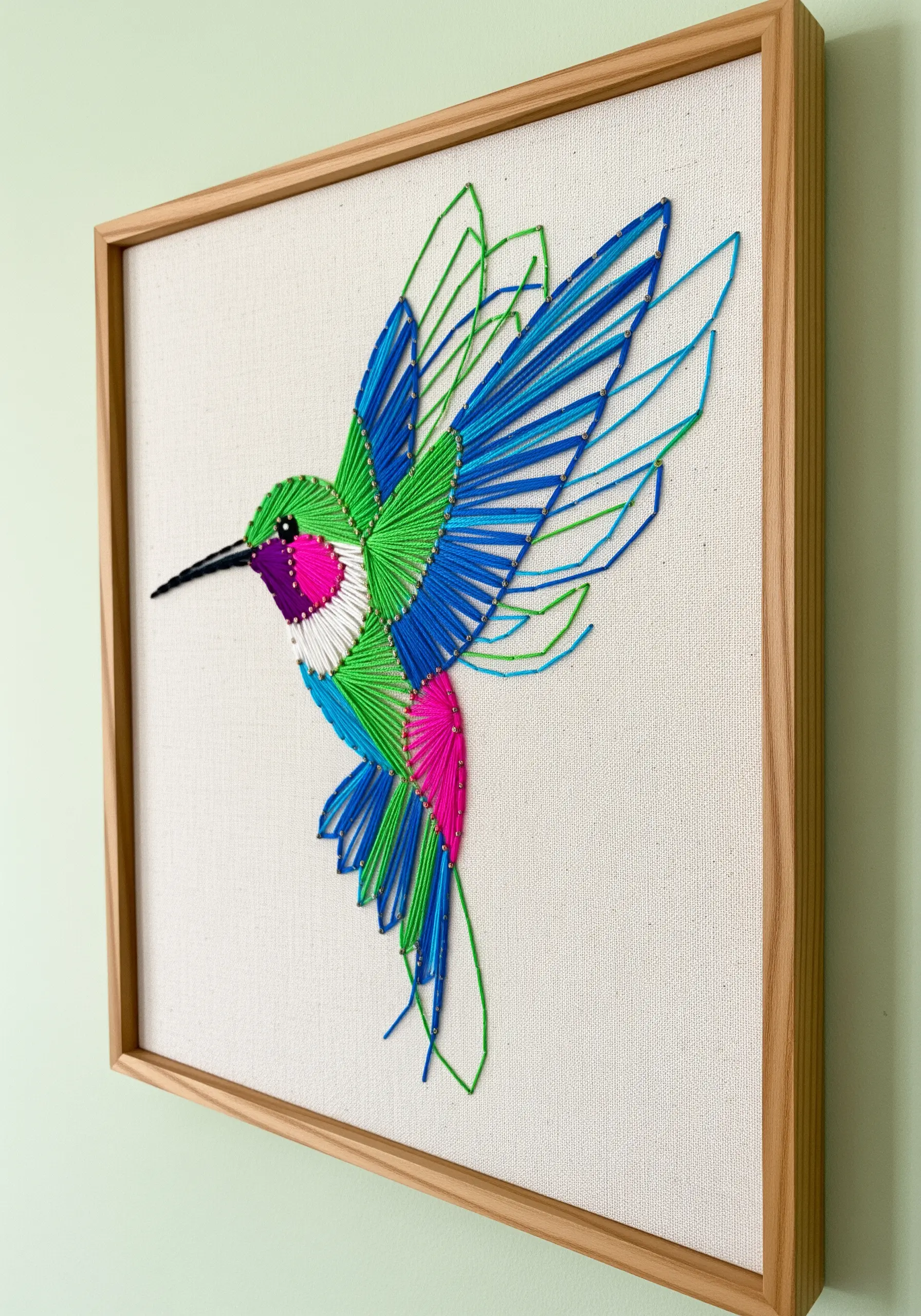 A vibrant string art hummingbird with blue, green, and pink color-blocked feathers.