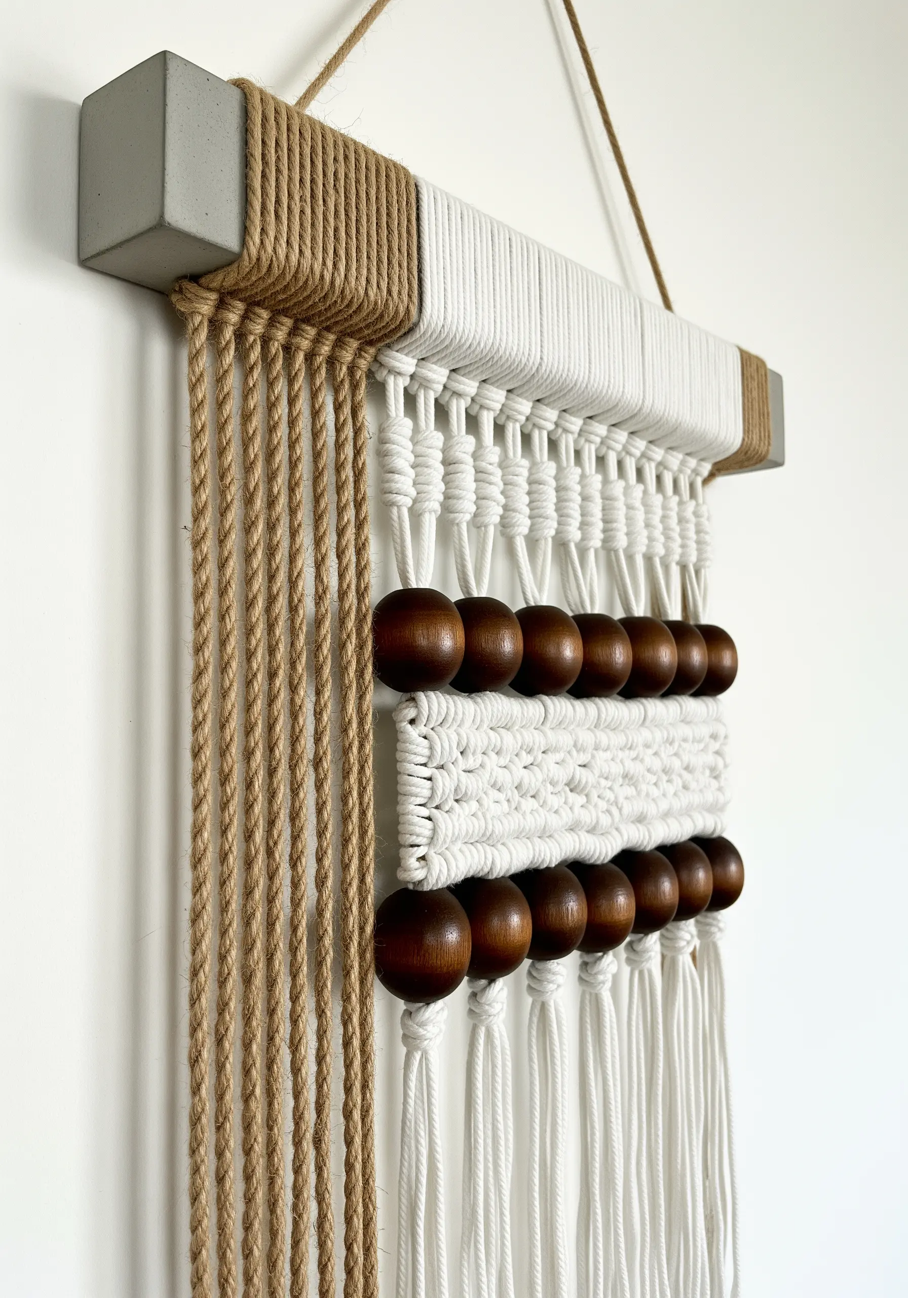 A modern macrame wall hanging with jute, cotton, and wood beads on a concrete bar.