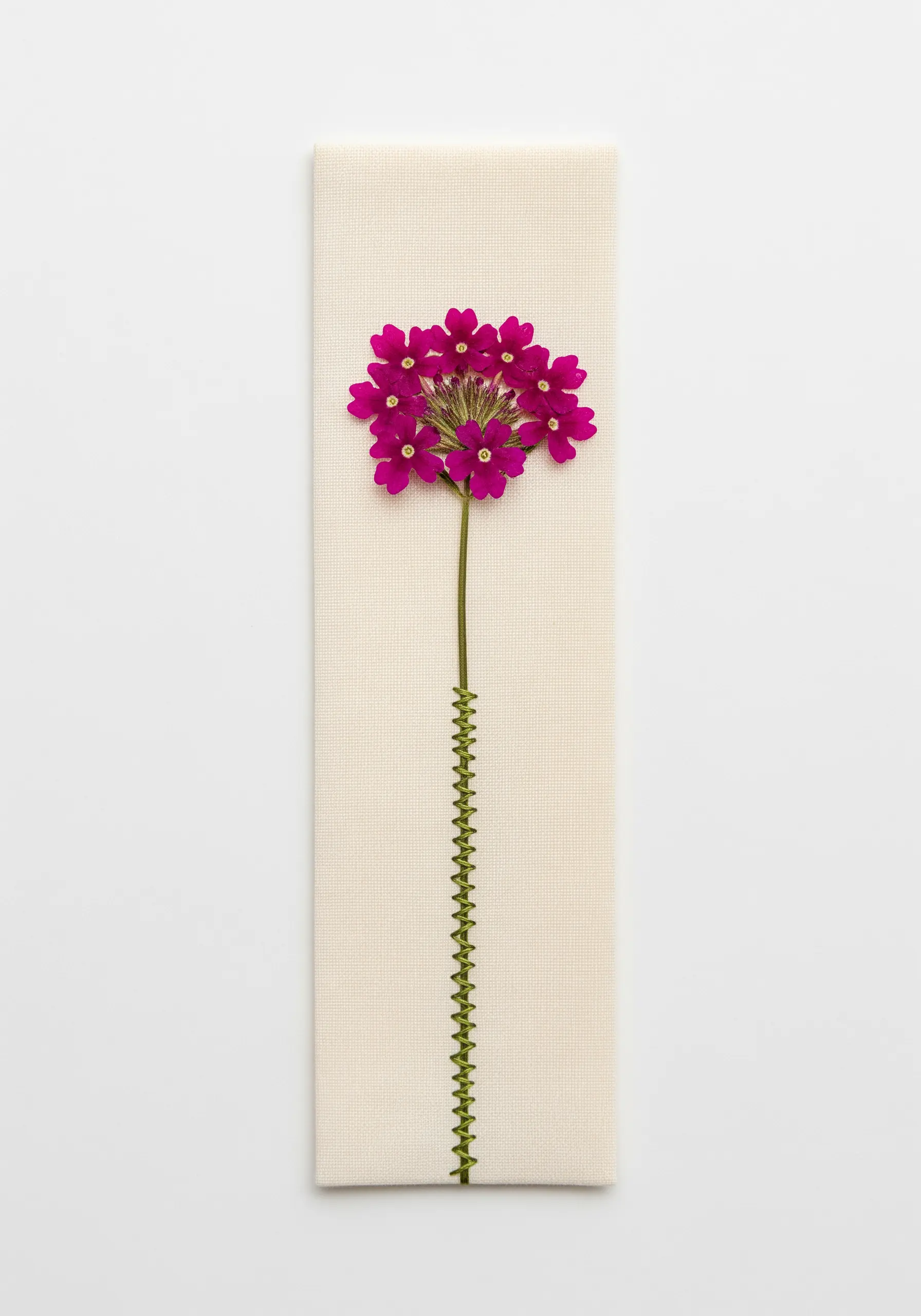 A single pressed magenta flower with a graphic green embroidered zigzag stem on a canvas bookmark.