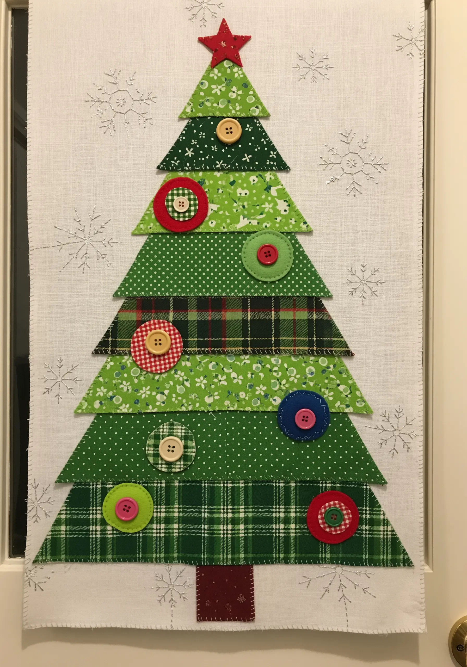 A fabric Christmas tree wall hanging made of layered green patterned fabrics and decorated with buttons.