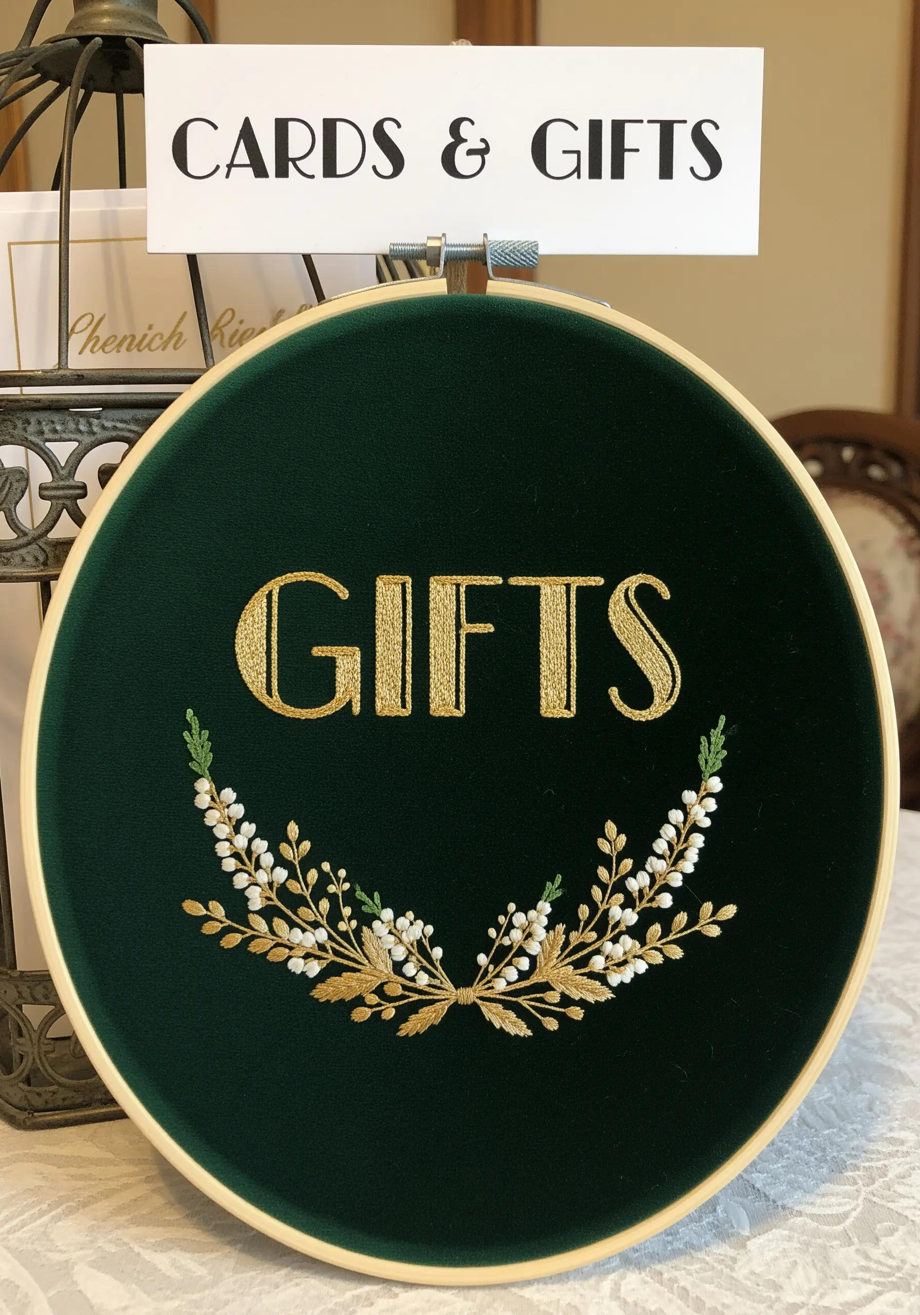 A dark green velvet hoop sign embroidered with 'Gifts' in gold thread and a delicate floral wreath.