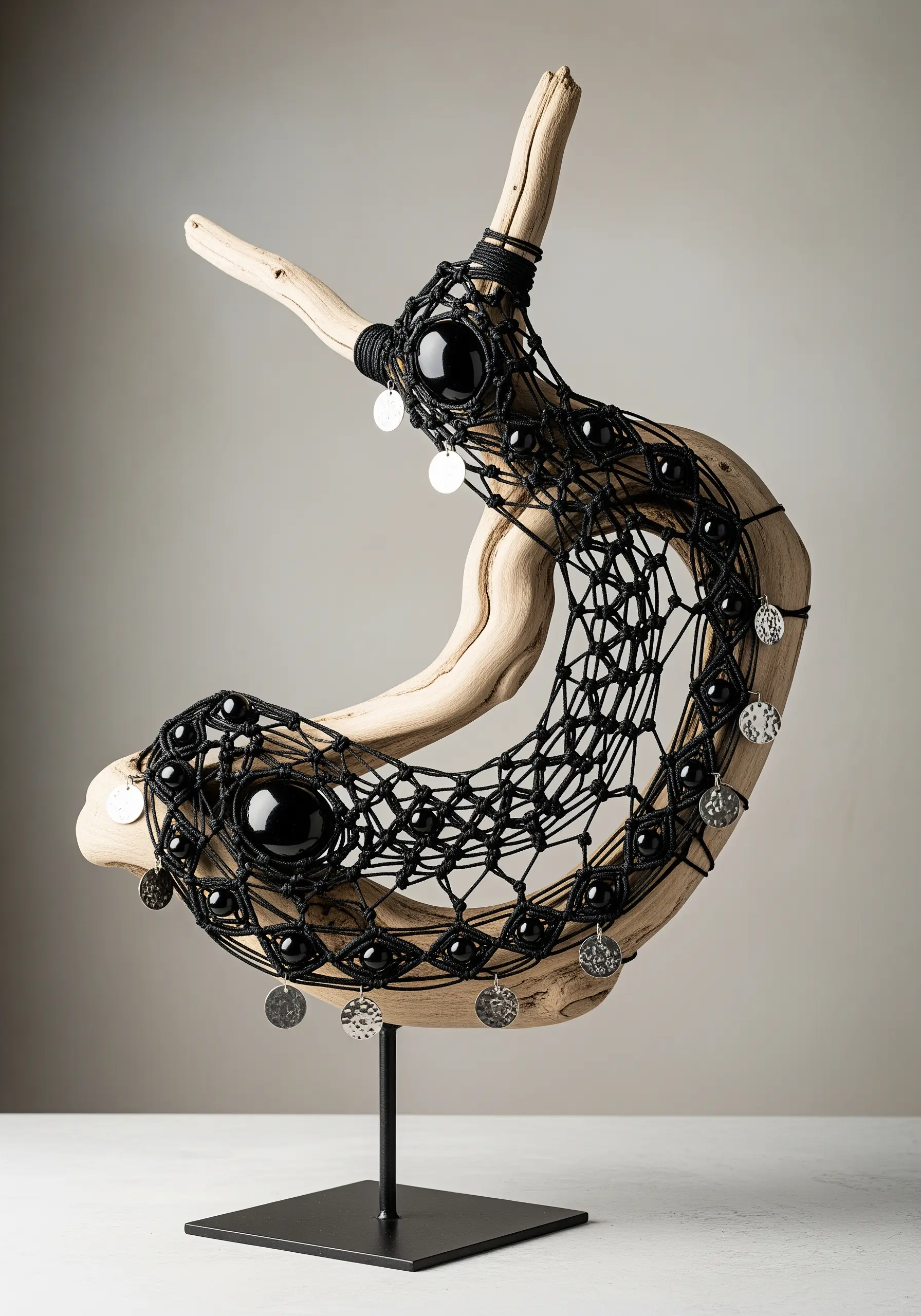 A piece of curved driftwood is wrapped in black macrame netting with large black beads and silver coin charms.