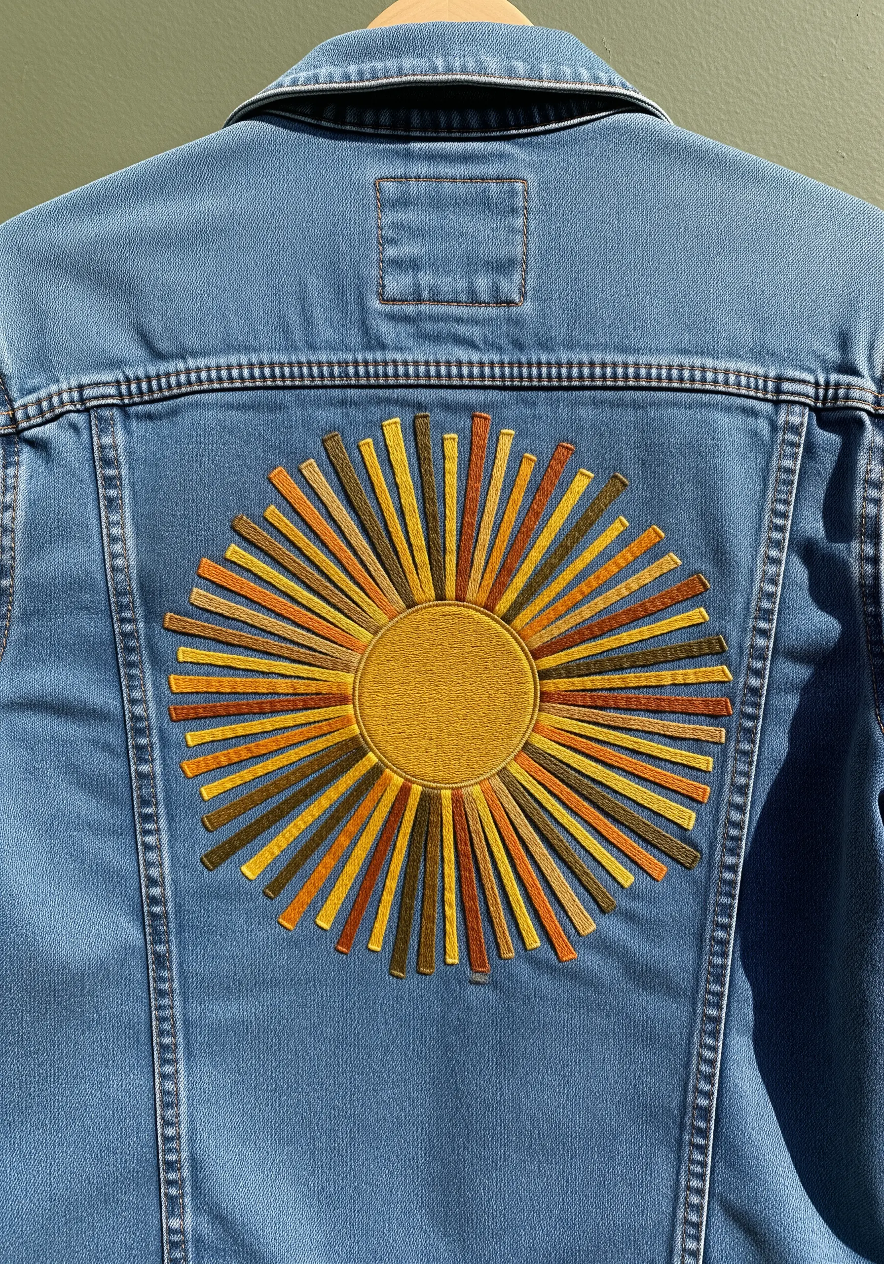 A large sunburst motif embroidered on the back of a denim jacket with satin stitch.