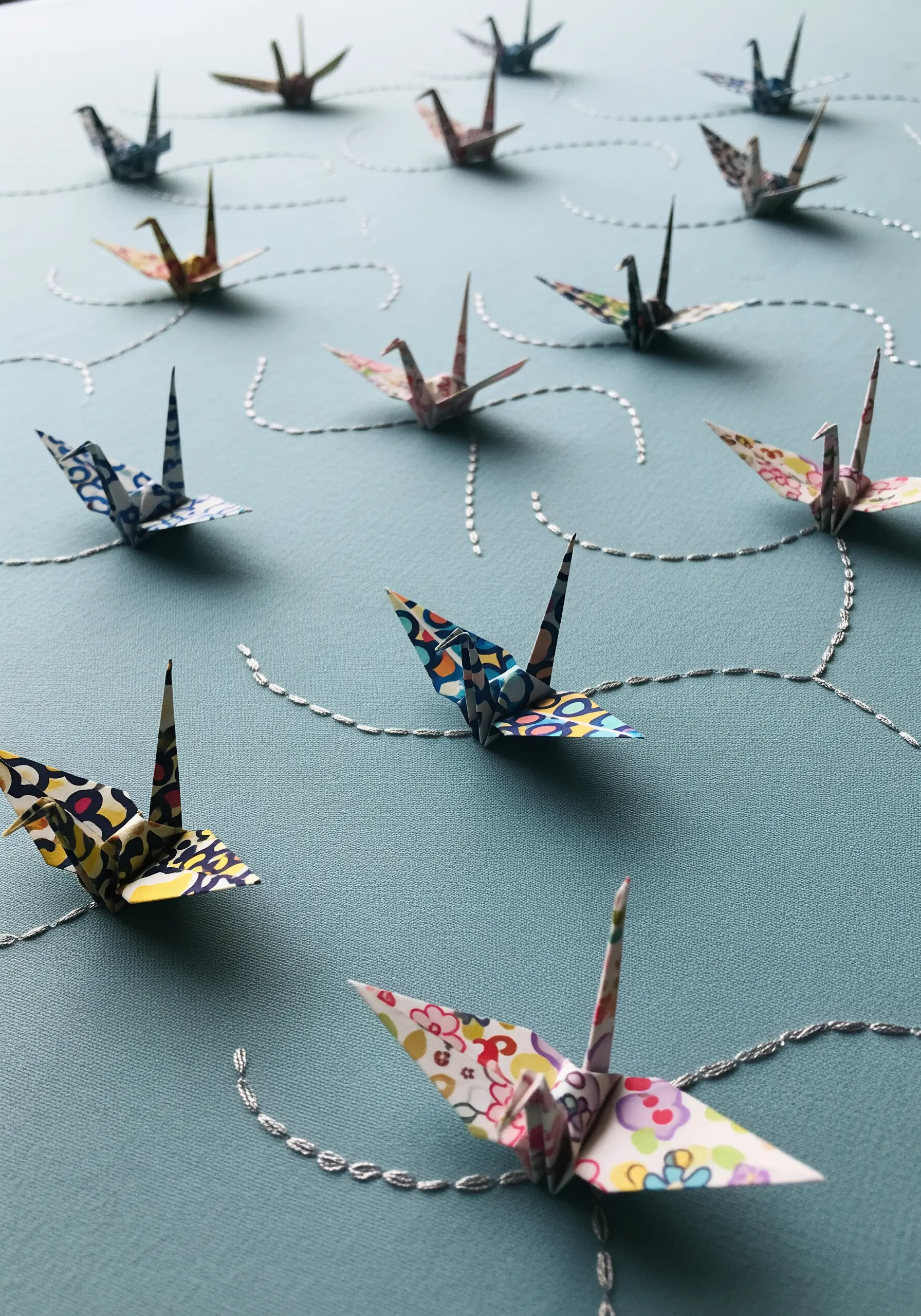 Small origami cranes arranged on blue fabric, connected by swirling paths of silver running stitch.