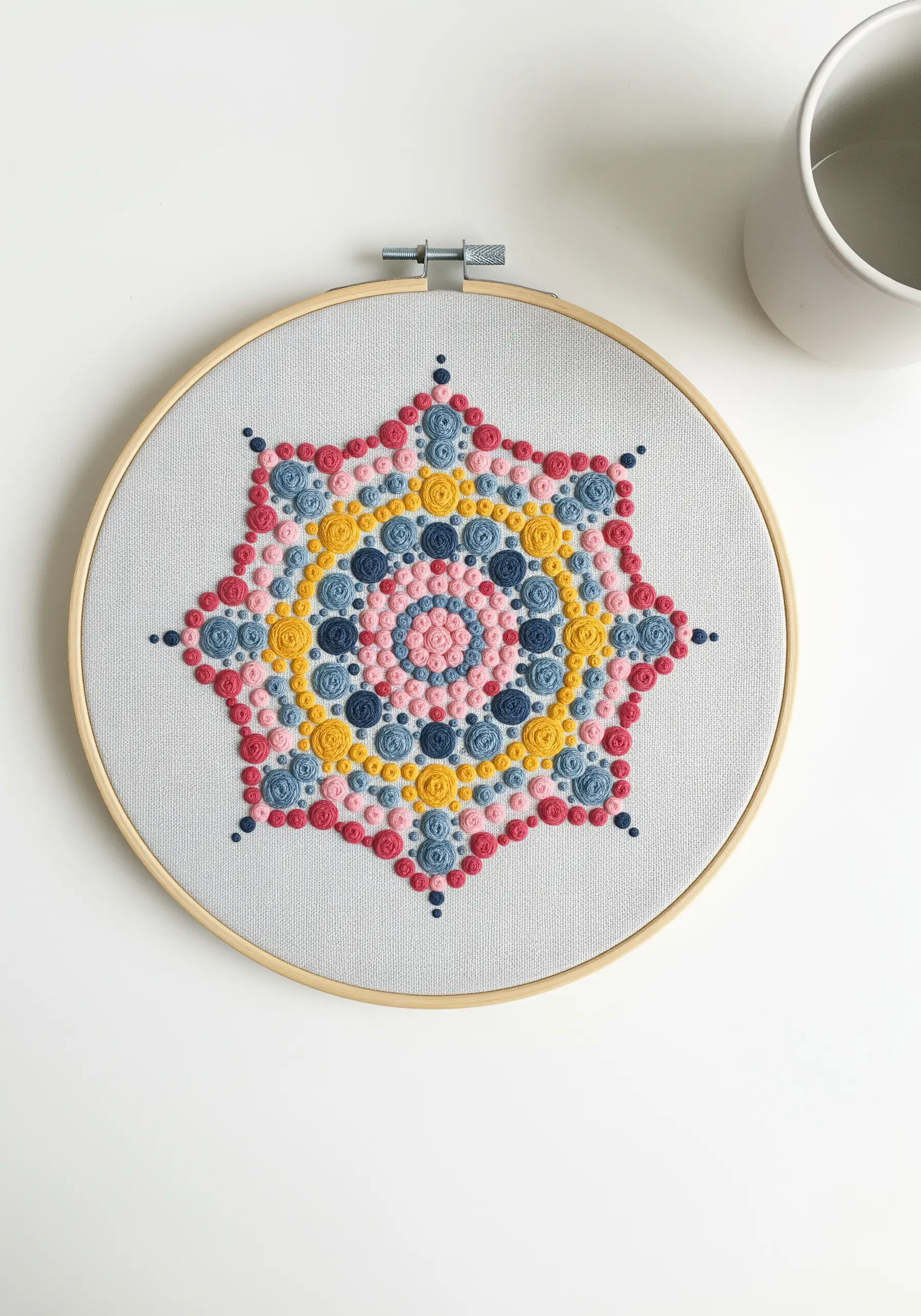 A geometric star mandala created entirely from colorful French knots in pink, blue, and yellow.