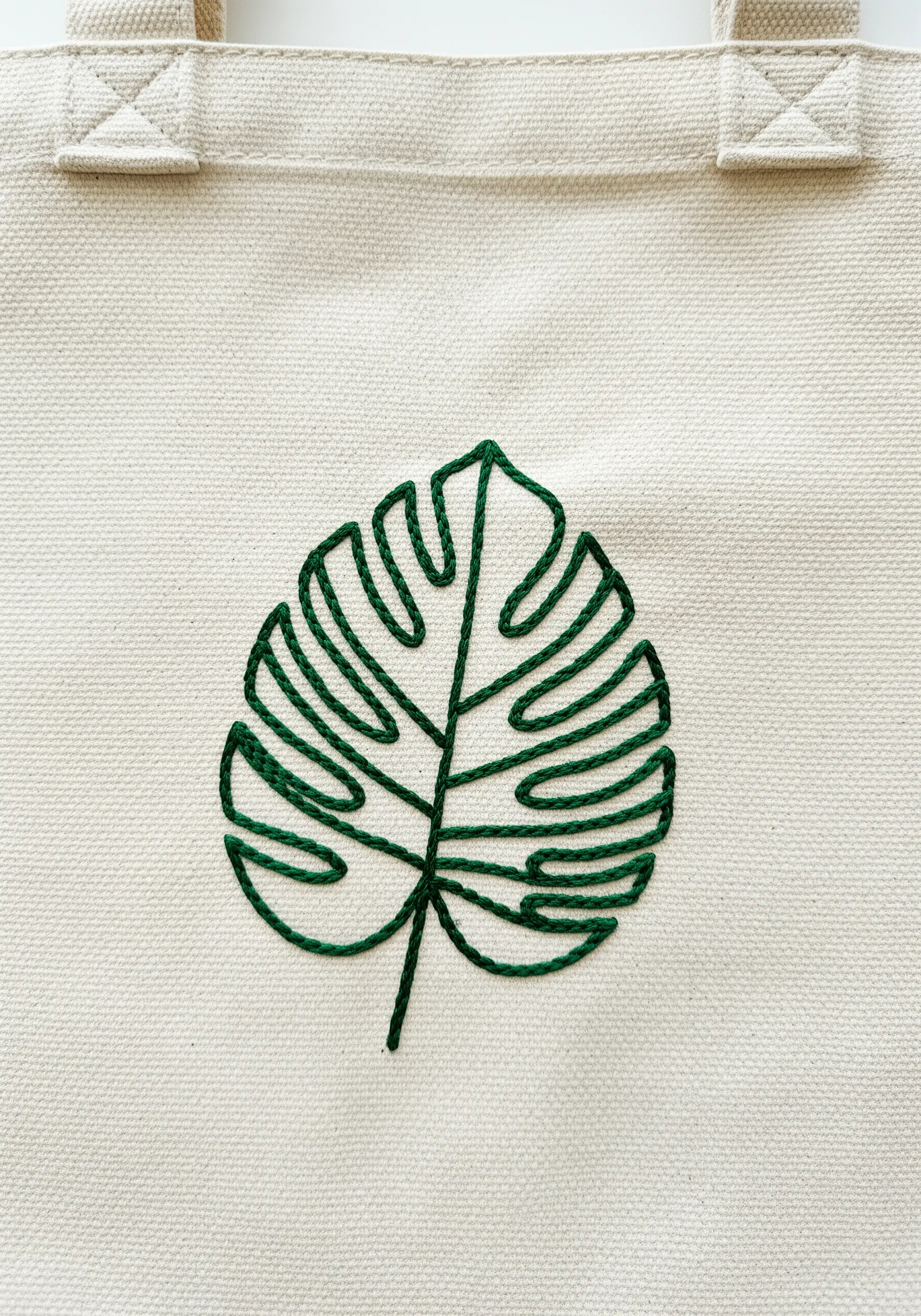 A line-art embroidery of a monstera leaf in dark green thread on a tote bag.