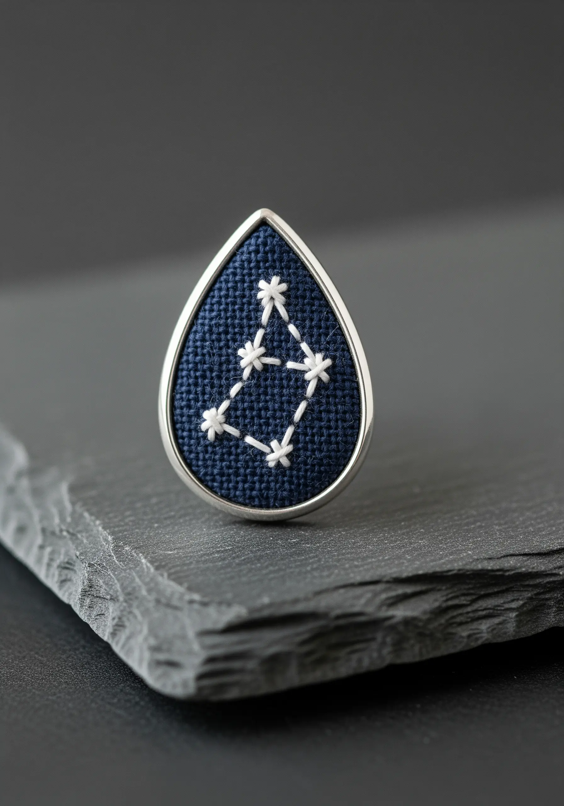 A white embroidered constellation on dark navy fabric set in a silver teardrop bezel.