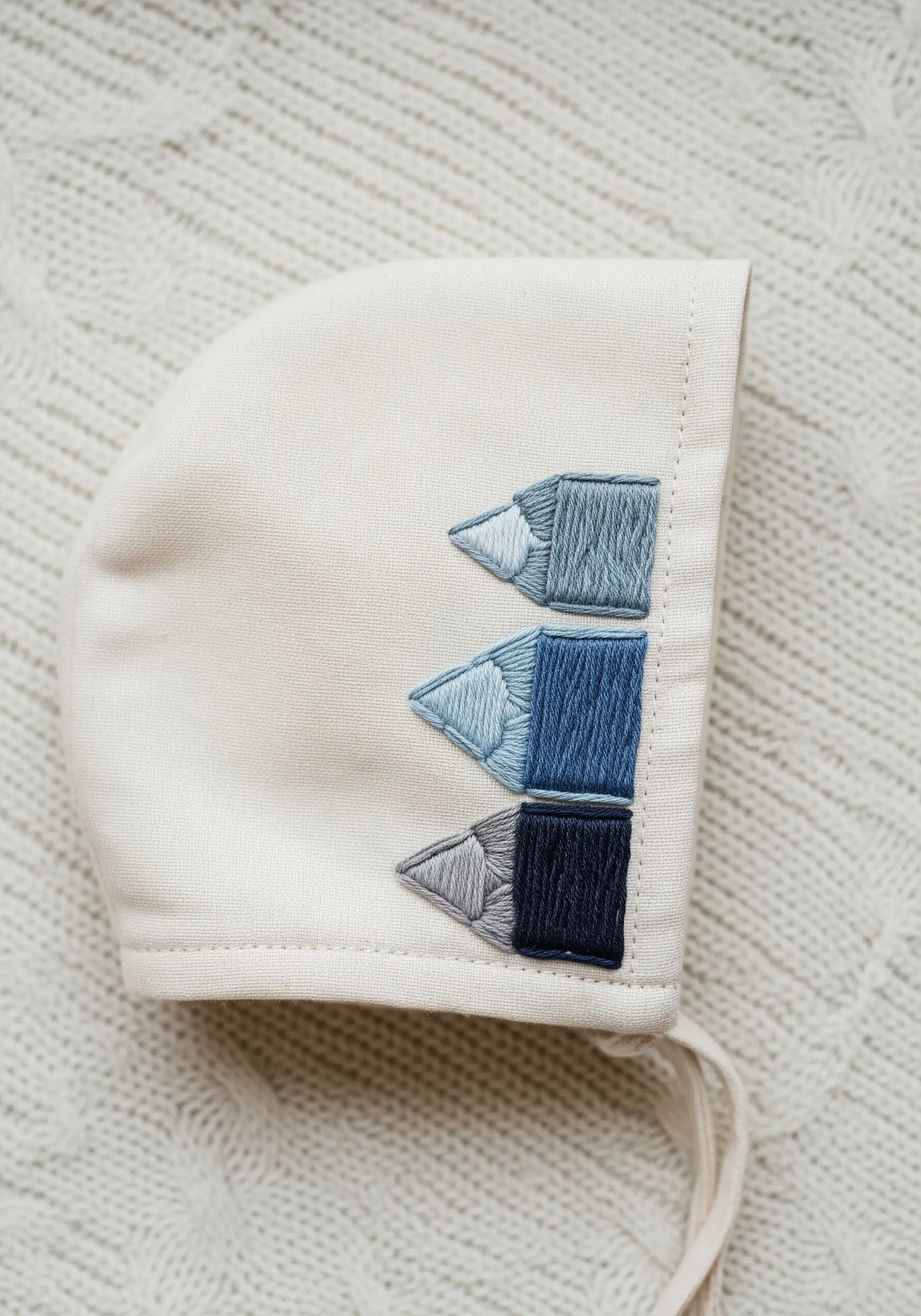 Three colored pencils embroidered on a bonnet using a blue color gradient.